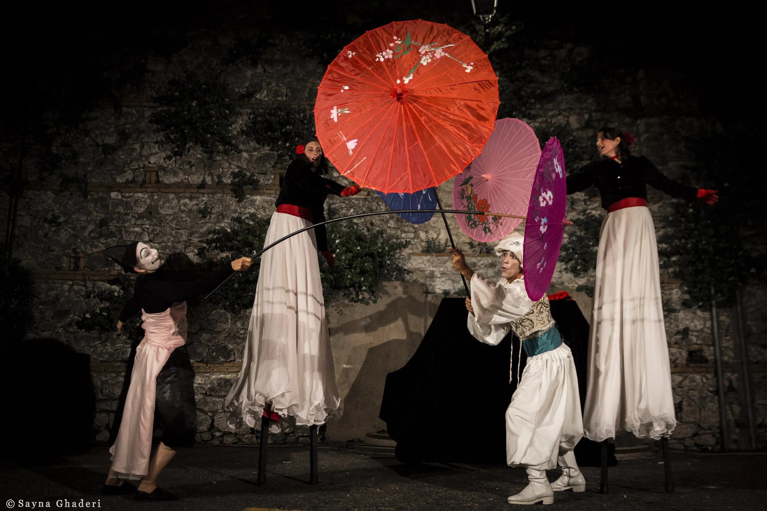 Personaggi in costume di teatro delle marionette con ombrelli colorati, in scena all'aperto di notte.