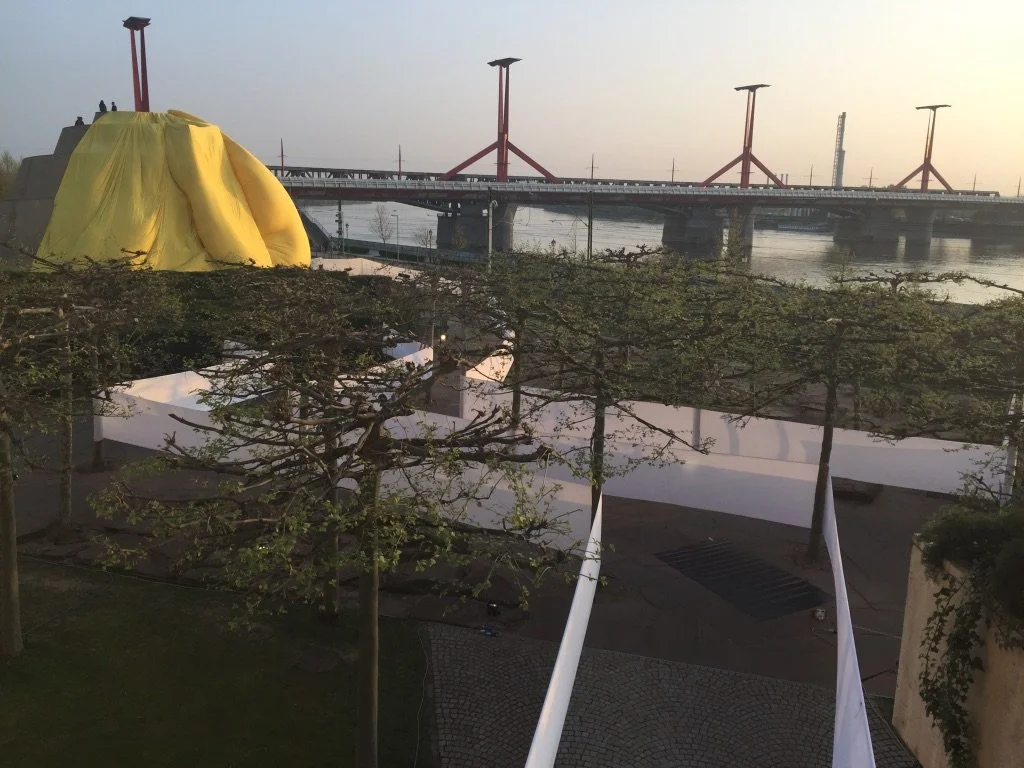 Struttura temporanea con tenda gialla e alberi, ponte sul fiume con strutture rosse sullo sfondo, al tramonto.