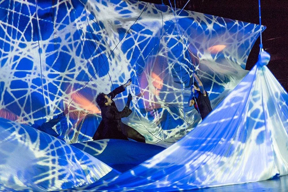 Persone coinvolte in un'installazione artistica o performance con tessuti blu e luci colorate, creando effetti di luce e ombra.