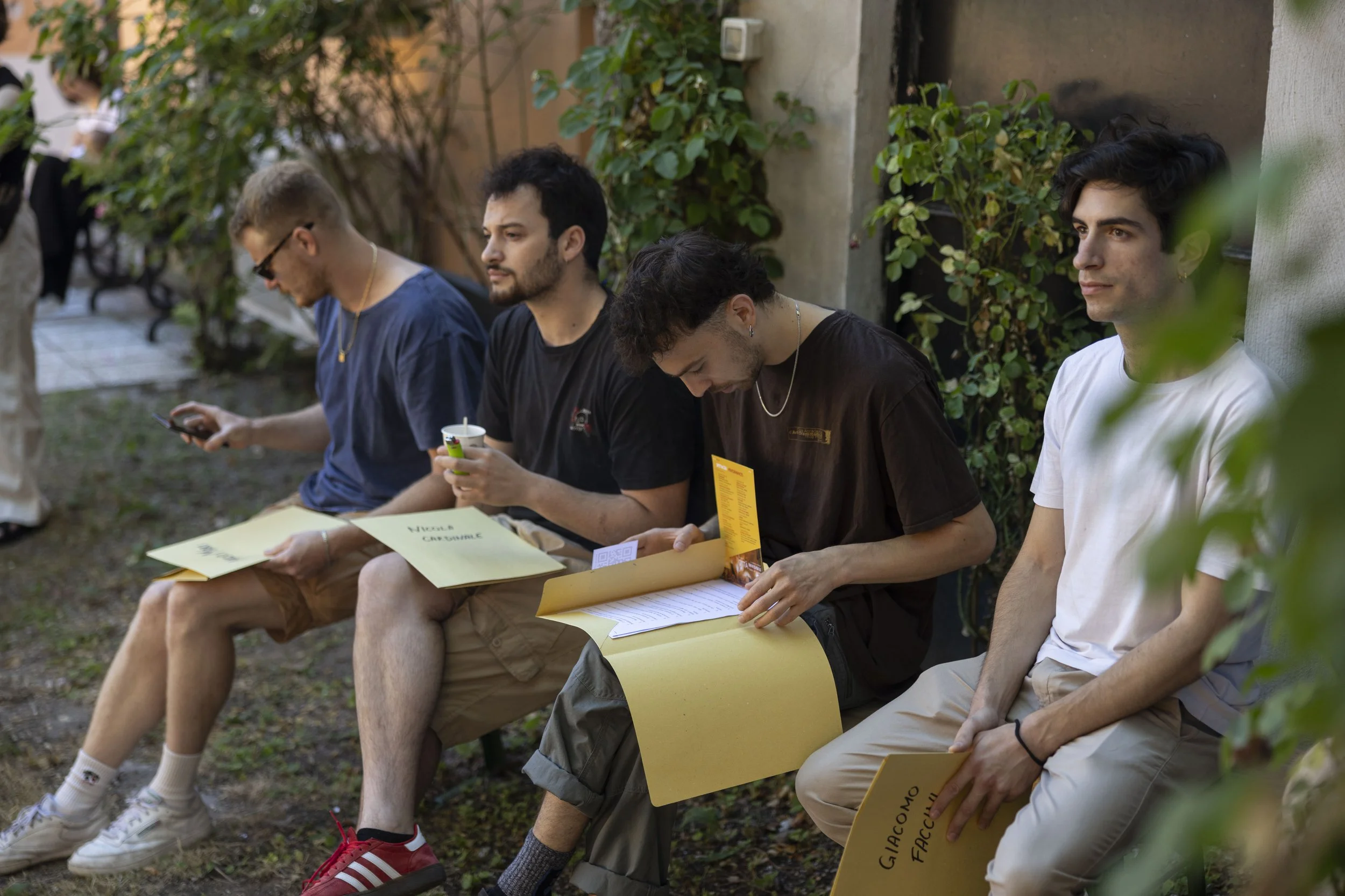 Cinque giovani seduti su una panchina all'aperto, alcuni leggendo documenti, altri usando il telefono, in un ambiente con piante e un muro.