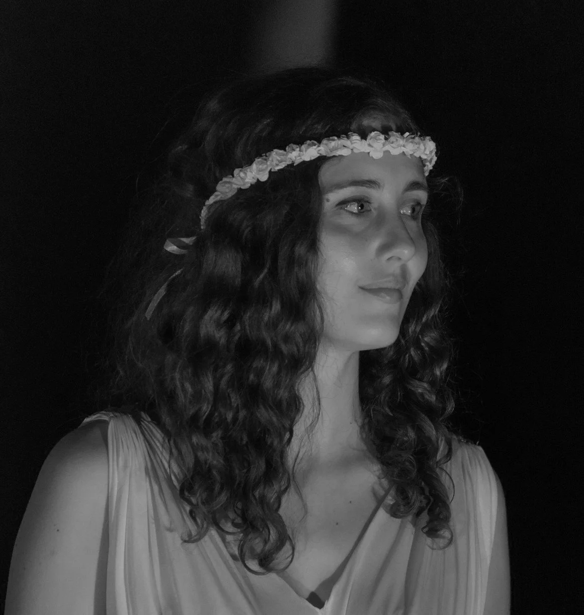 Ragazza con capelli ricci, fasciata con corona di fiori, in abito chiaro, vista di profilo, sfondo scuro.