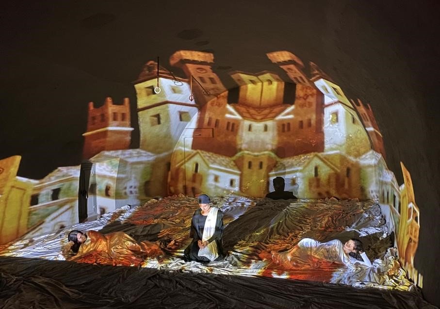 Una scena teatrale con persone sdraiate e in piedi, con un castello e edifici storici proiettati sullo sfondo e sul palco