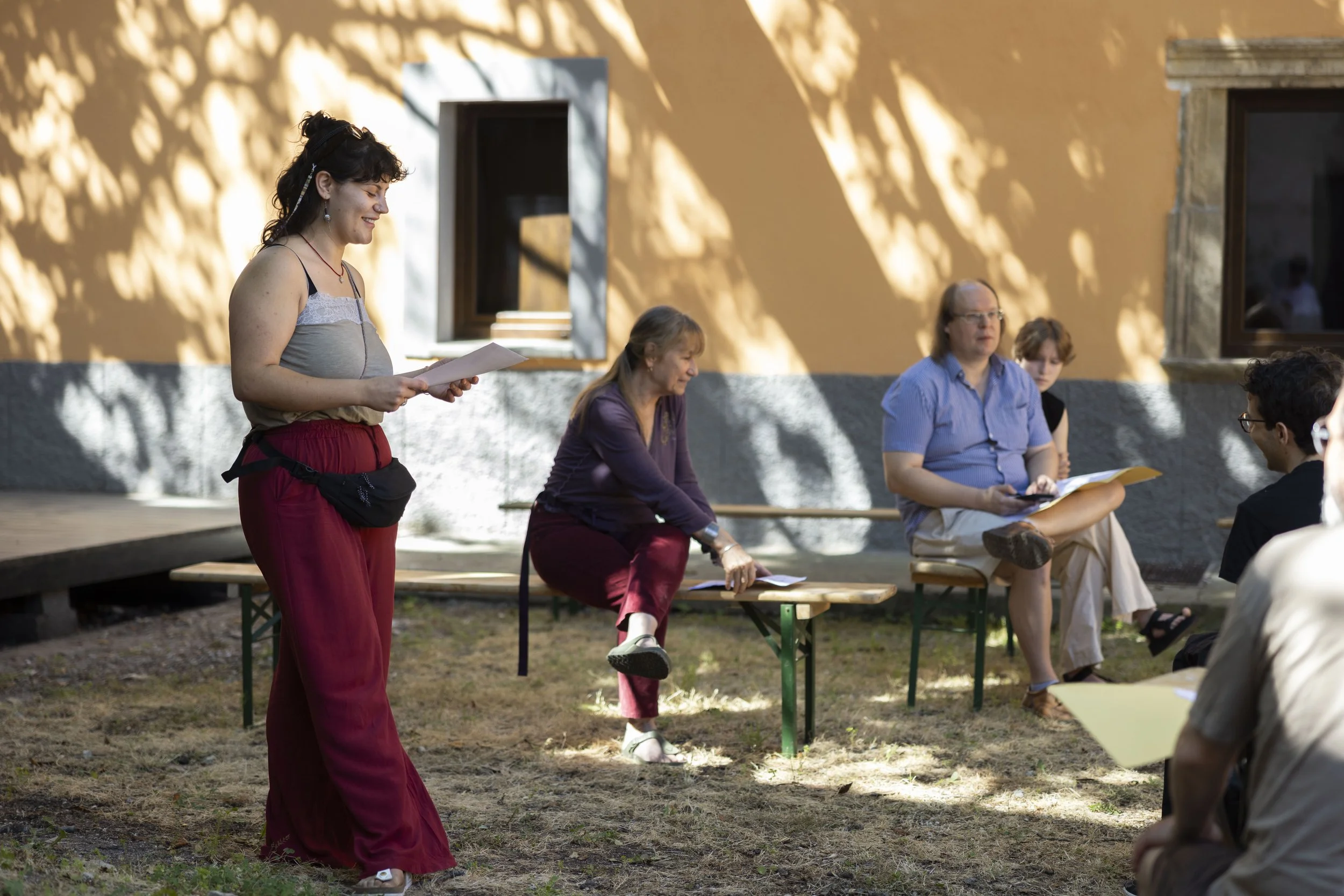 Gruppo di persone che partecipano a una discussione all'aperto, con alcune sedute su panche e una donna in piedi che legge da un foglio, in una zona ombreggiata con pareti colorate.