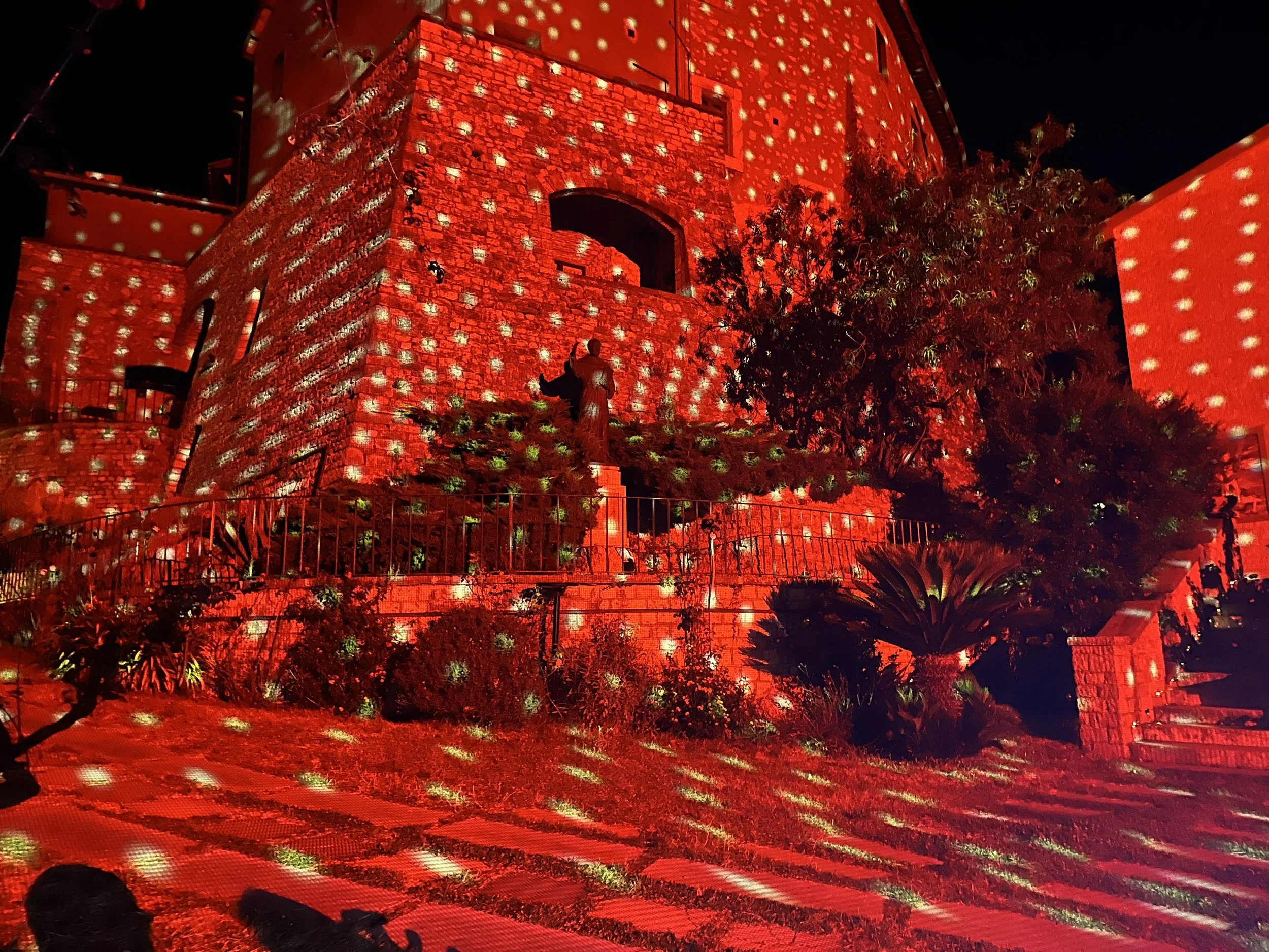 Edificio reso rosso con luci e proiezioni, decorato con alberi e piante nel giardino di fronte.