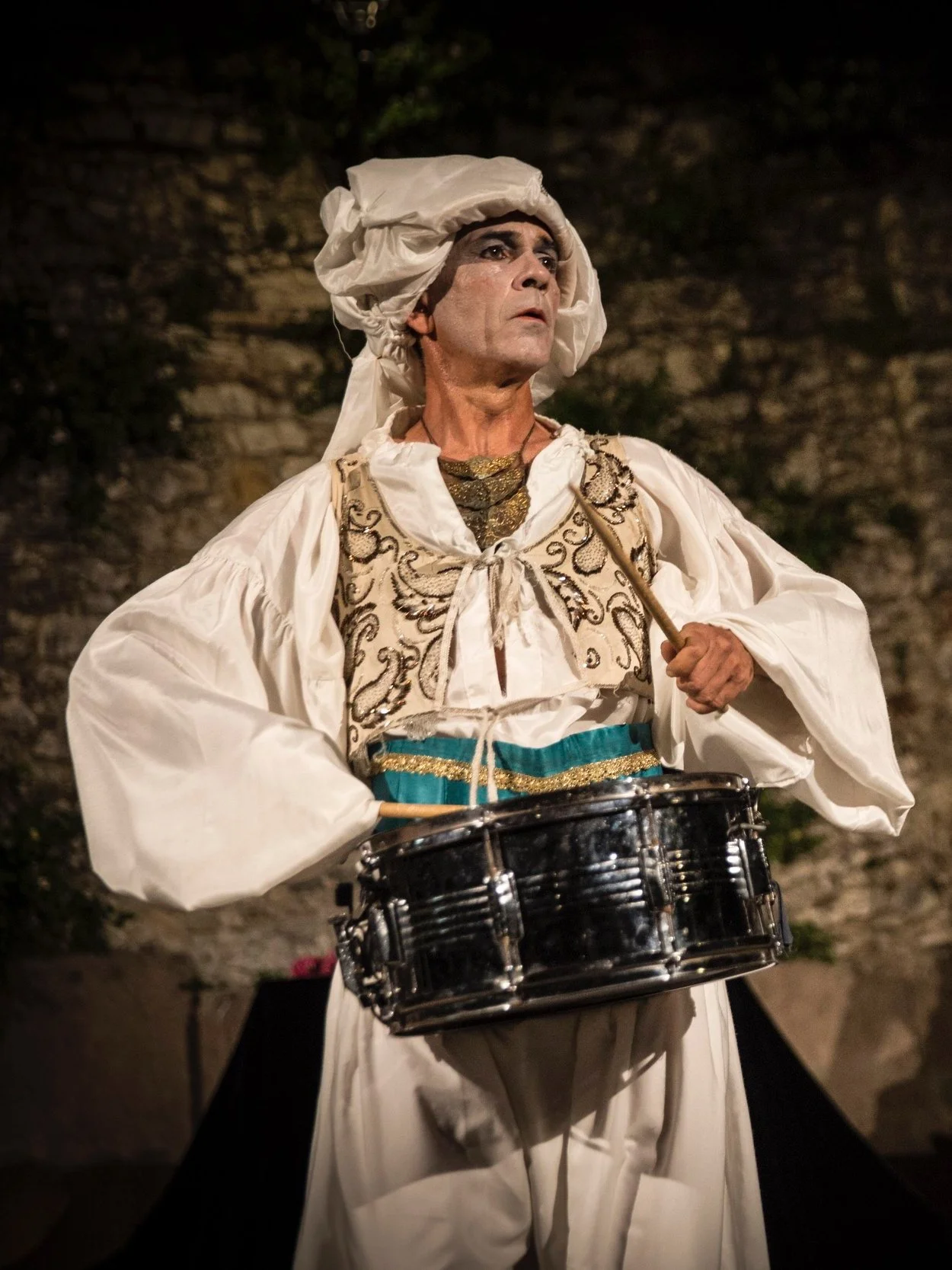 Persona in abiti storici con cappello e vestiti elaborati, che suona un tamburo in un contesto esterno notturno.