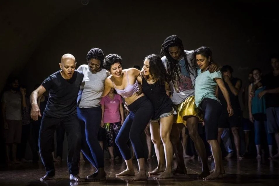 Un gruppo di persone in piedi in cerchio, che si tengono per le spalle e sorridono mentre ballano in un ambiente interno con luci soffuse.