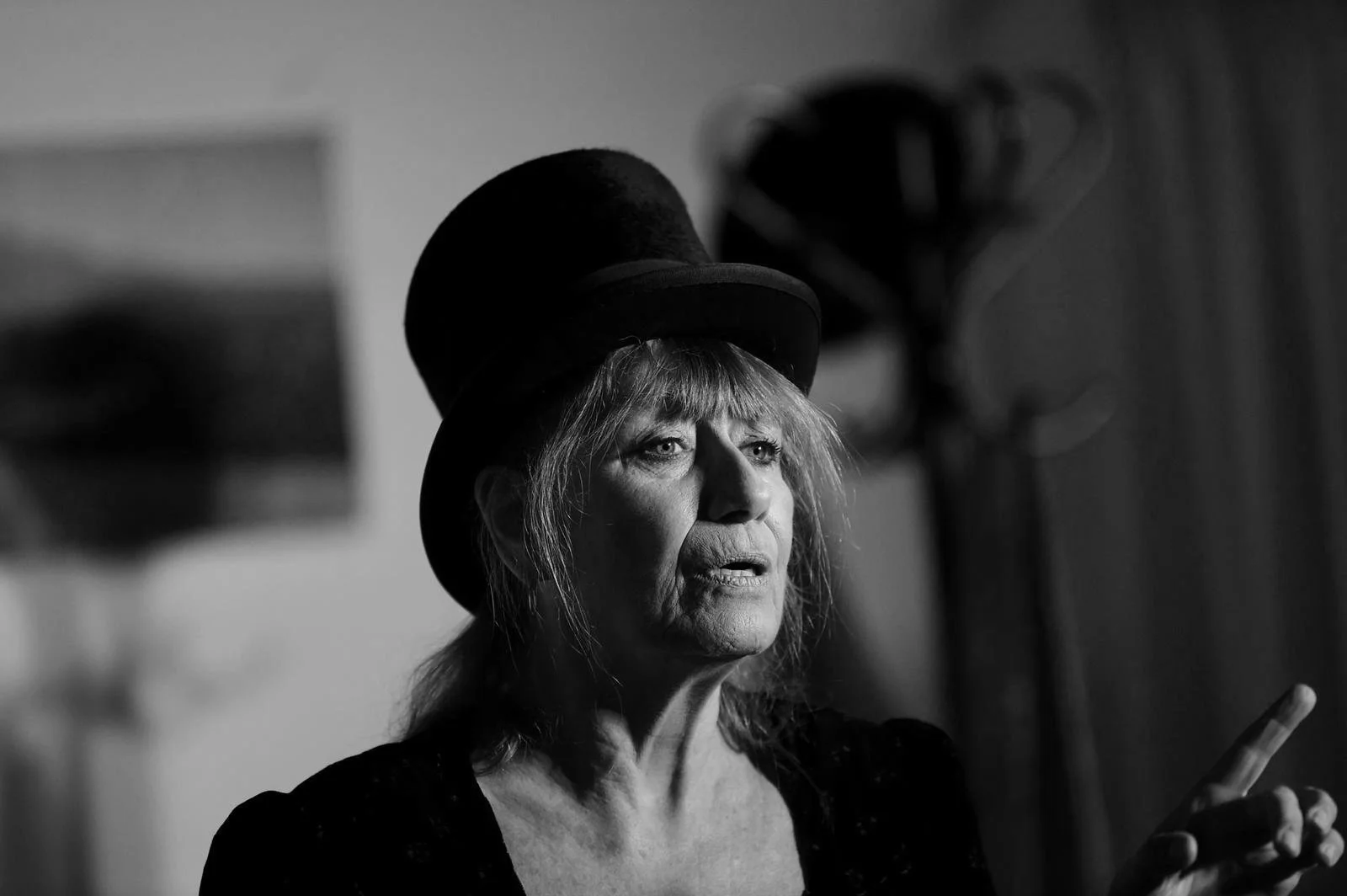 Donna anziana con cappello nero, capelli grigi disordinati, espressione sorpresa o preoccupata, che tiene in mano uno smartphone, sfondo sfocato.