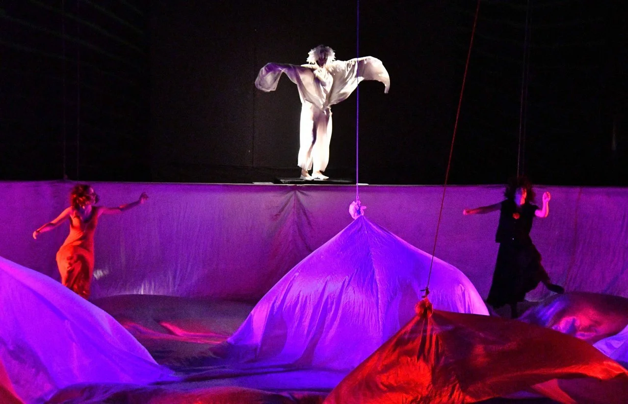 Performance teatrale con tre attori su scena, uno in alto con braccia aperte e in costume bianco, e due in basso, una vestita di rosso l'altra di nero, circondati da drappi o teli colorati illuminati da luci rosa e viola.