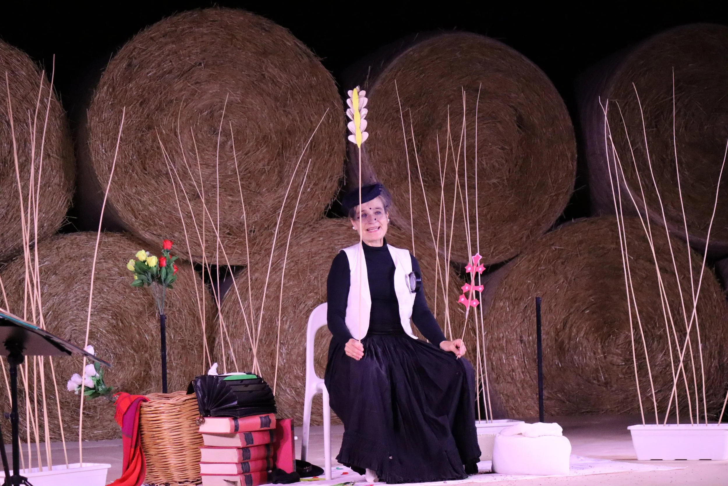 Una donna vestita di nero e bianco, seduta su una sedia bianca con sfondo di balle di fieno, circondata da fiori e scaffali con libri, durante una rappresentazione teatrale.