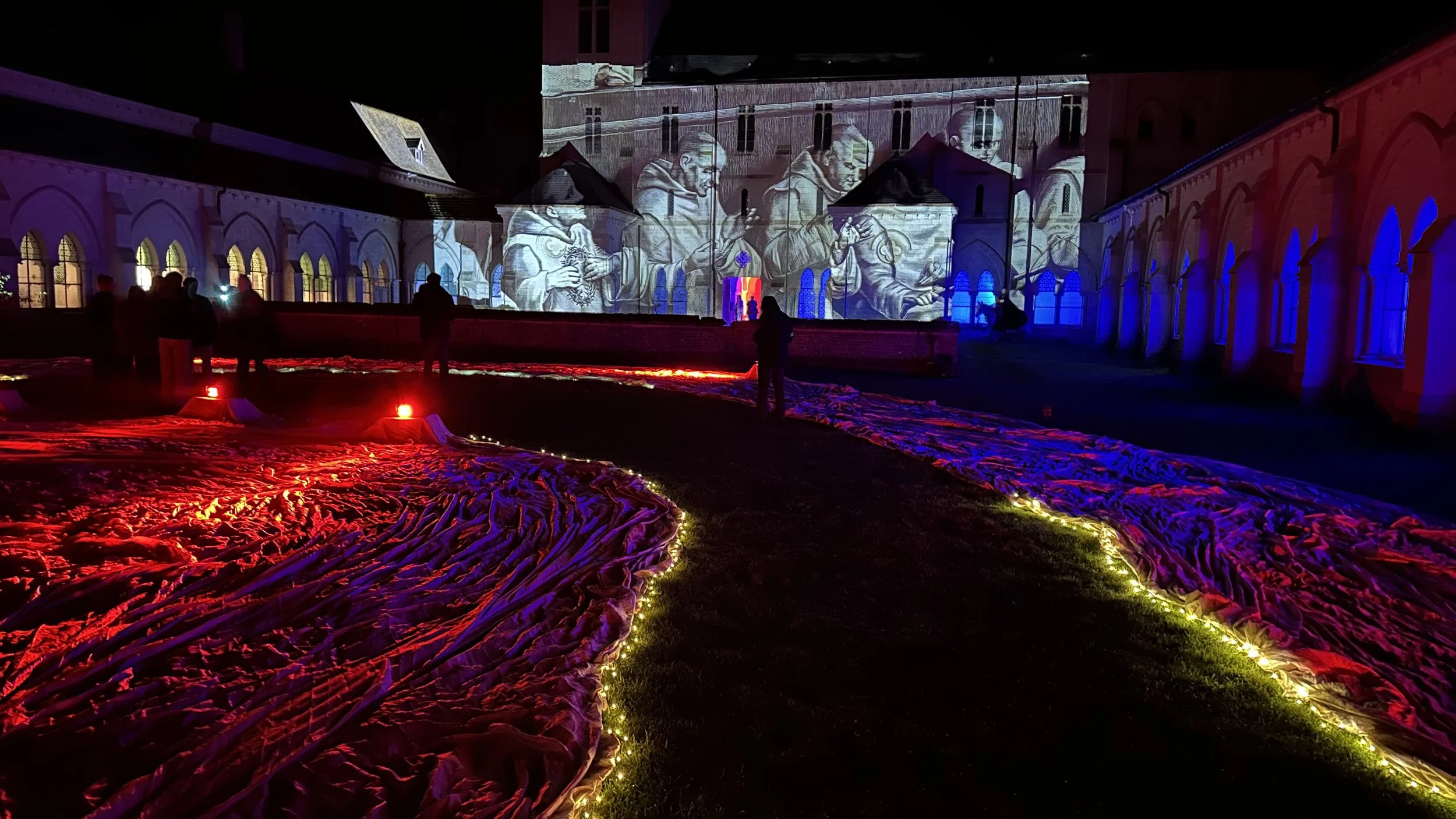 Installazione artistica con luci colorate, drappeggi di tessuto illuminato e proiezioni di dipinti storici su un edificio antico, durante una mostra notturna.