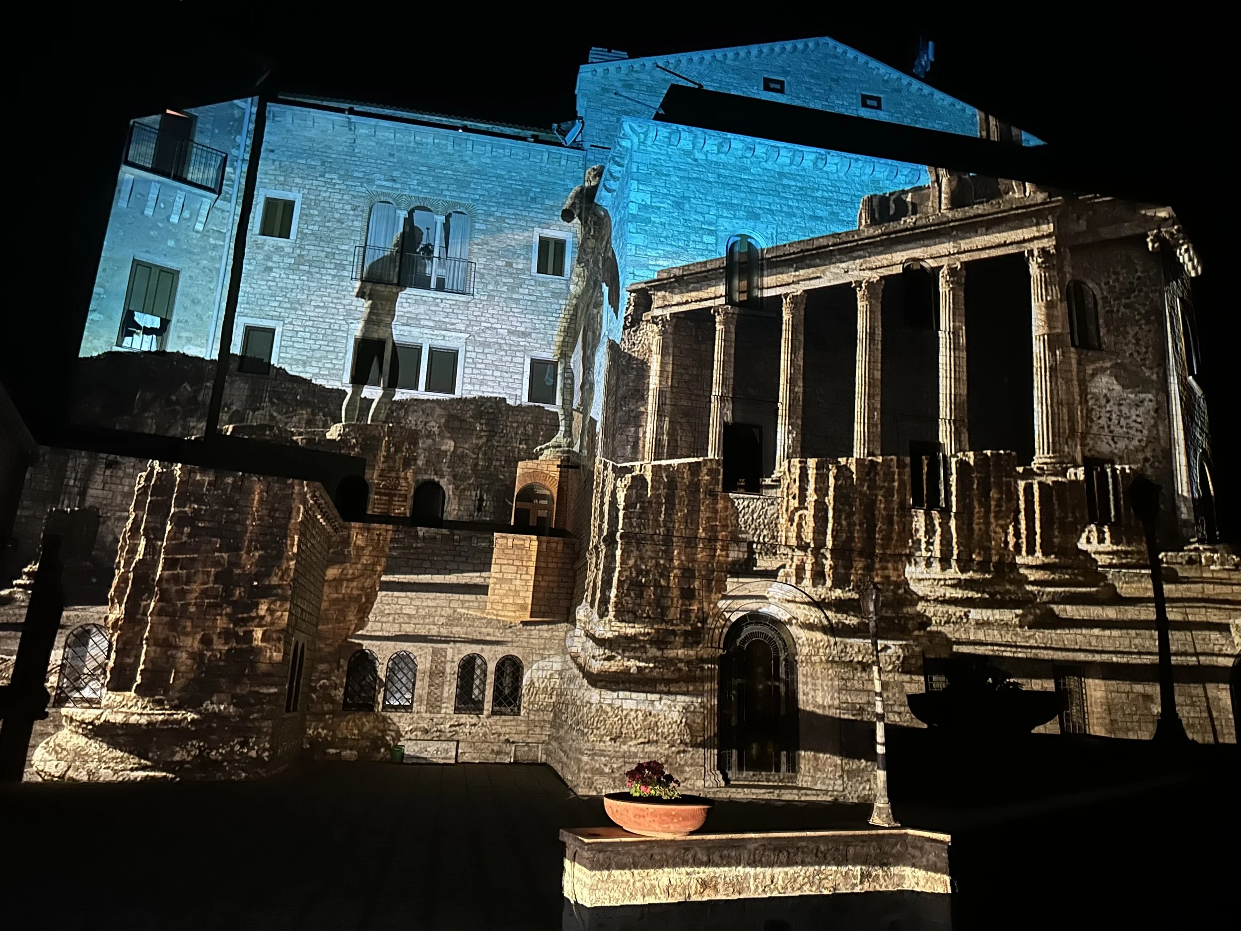 Proiezione di un'antica architettura su una parete con luci colorate, raffigurante un edificio storico con colonne e dettagli architettonici, con una pianta di fiori in primo piano.