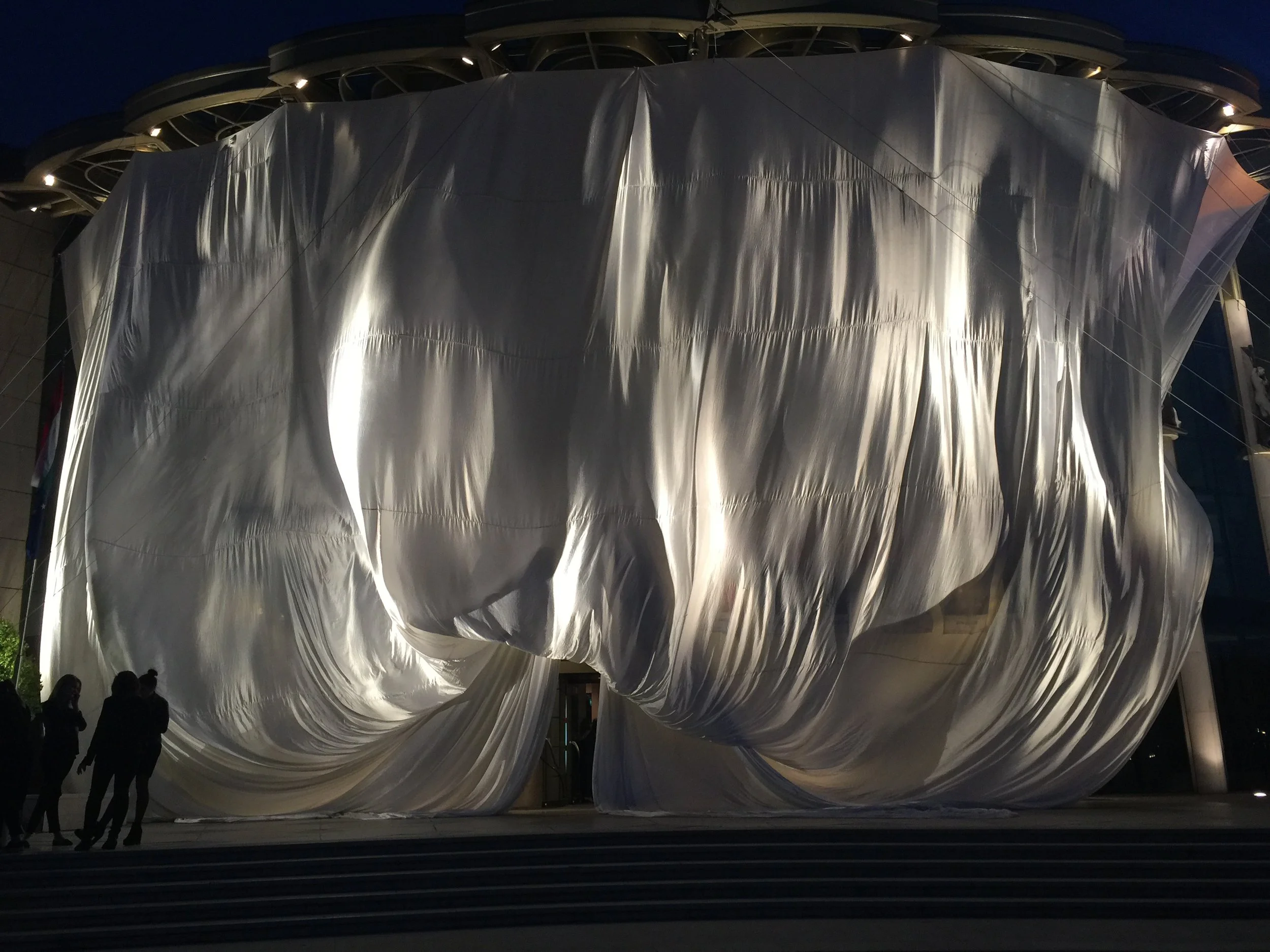 Edificio coperto da una grande tela bianca, illuminato di notte, con alcune persone in piedi davanti.
