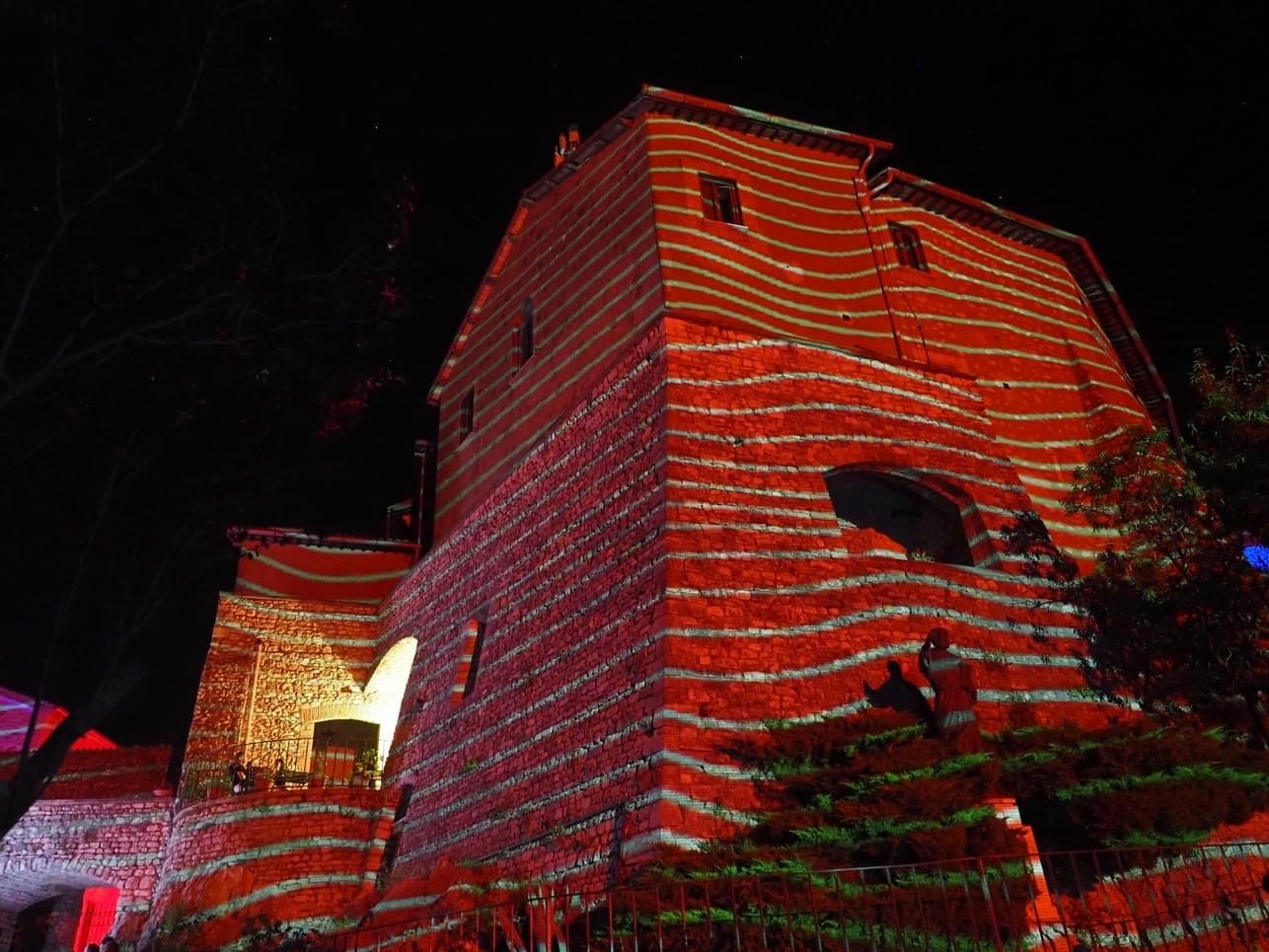 Castello illuminato con luci rosse e linee di luce verticali, di notte, con alberi intorno.