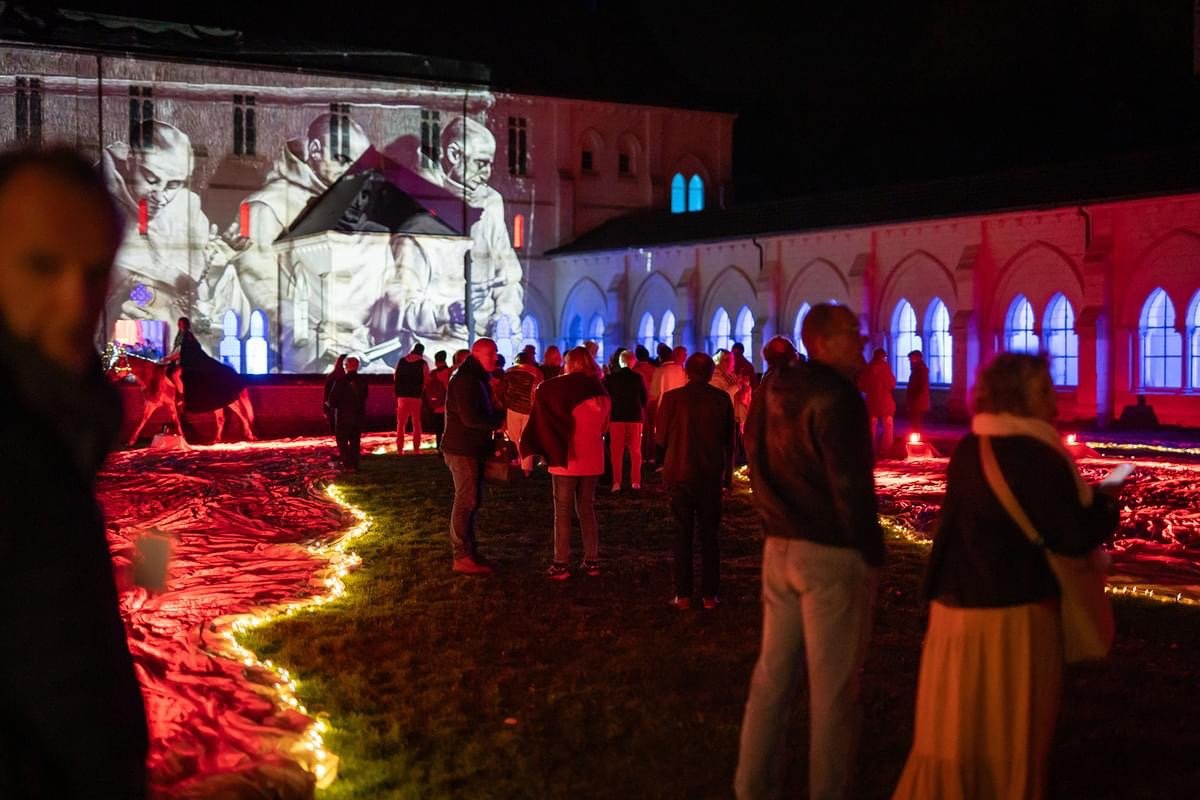 Festival notturno con proiezioni di immagini su un edificio storico, persone che guardano e si godono lo spettacolo, luci colorate e un percorso luminoso sull'erba.