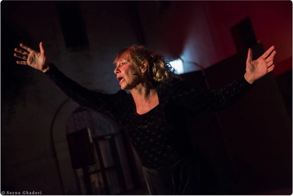 Una donna anziana con capelli riccioli castani si esibisce in una performance teatrale, con le braccia aperte e un'espressione emotiva, in un ambiente teatrale con luci soffuse.