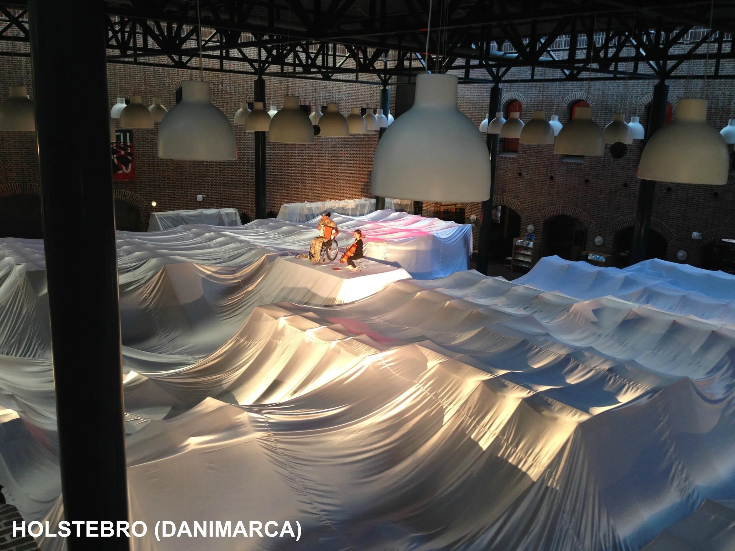 Interno di un edificio con soffitto di travi in ferro e grandi lampadari bianchi. Sul pavimento ci sono forme che sembrano onde di stoffa o plastica, con due sculture o modellini di persone al centro.
