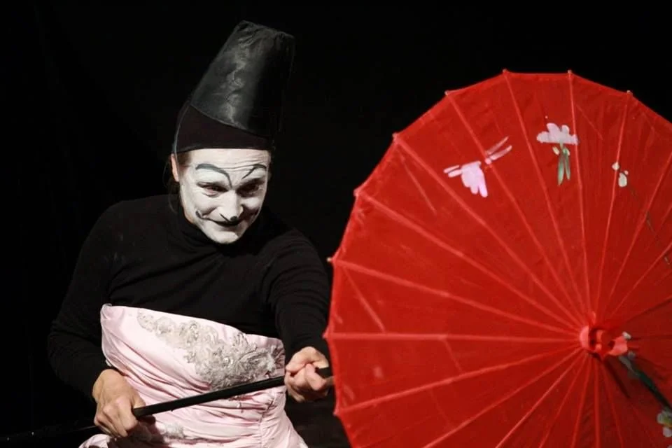 Artista con trucco da mimo e cappello nero tiene un ombrello rosso decorato con farfalle e fiori, sullo sfondo scuro.