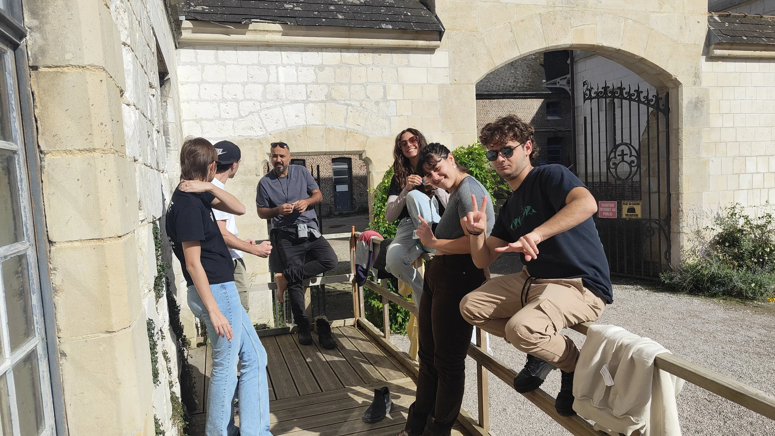 Gruppo di giovani che si rilassano e socializzano su un ponte di legno di fronte a un edificio di pietra, alcuni fanno segni con le mani, altri si abbracciano, in un ambiente esterno soleggiato.