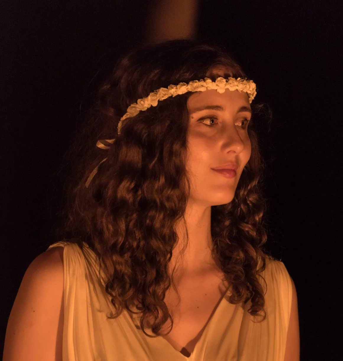 Donna con lunghi capelli ricci, corona di fiori, abito giallo chiaro, in un ambiente con illuminazione calda, sfondo nero.