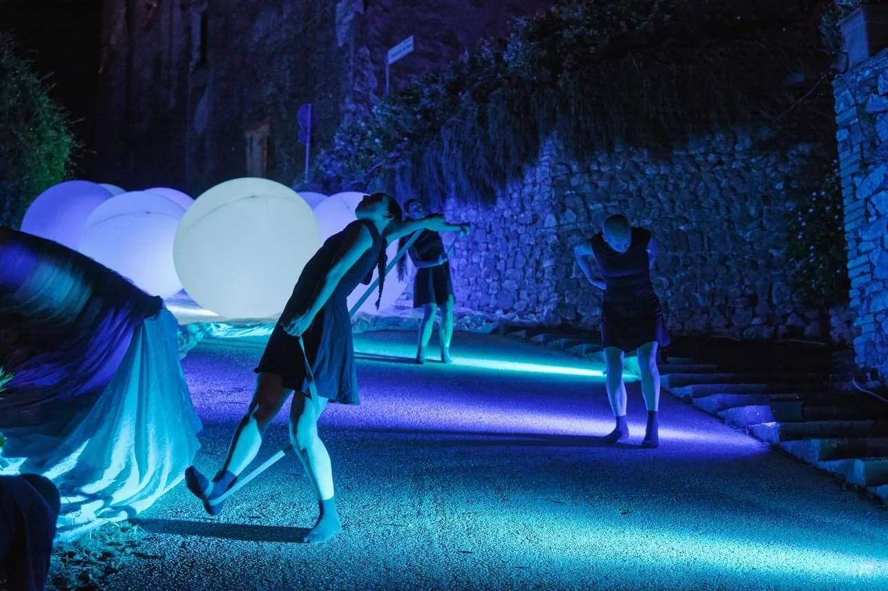 Persone in un'installazione artistica illuminata da luci blu e viola, con grandi sfere luminose in background, in un ambiente scuro con pareti di pietra.