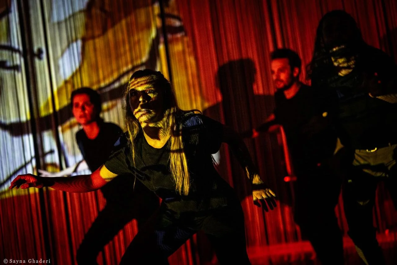 Tre persone che eseguono una performance teatrale o di danza in un ambiente con luci rosse e con proiezioni sullo sfondo, creando un'atmosfera drammatica.