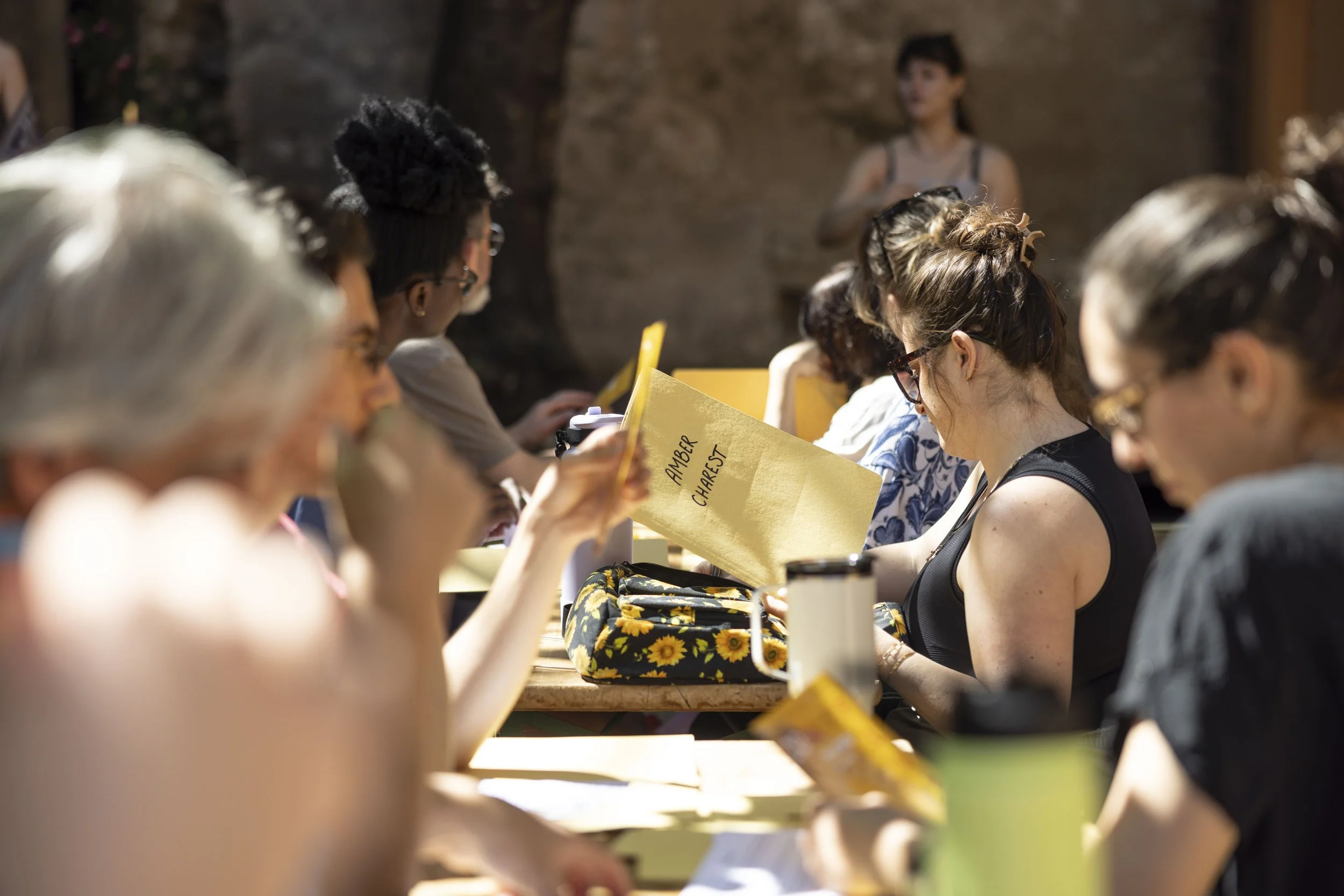 Gruppo di persone sedute a un tavolo, alcune leggendo fogli; sfondo con parete di pietra, luce naturale e ambiente all'aperto