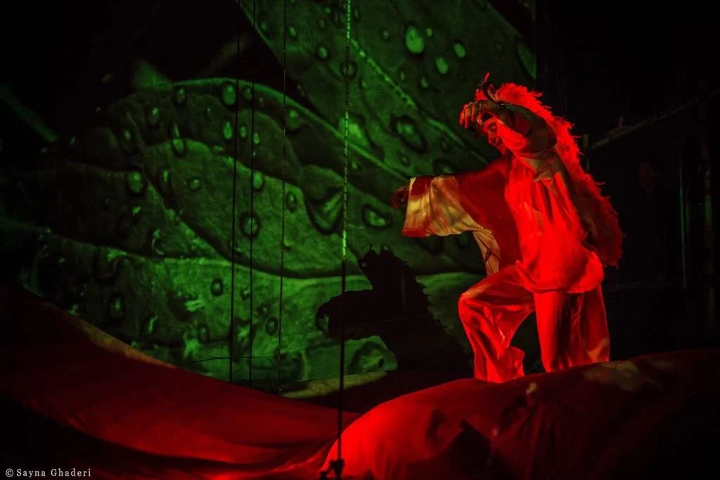 Una persona vestita con abiti rossi e una maschera da cavallo, che si trova su un palco con uno sfondo di luci verdi che raffigurano una foglia o una scena naturale.