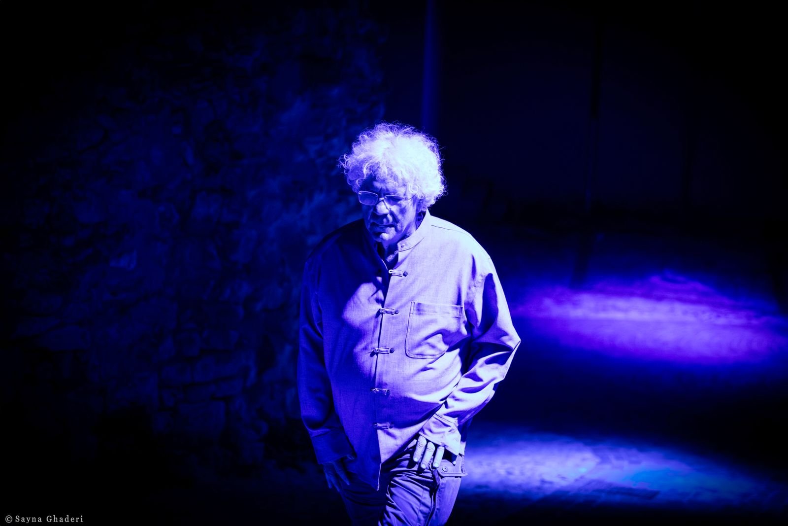 Uomo con capelli ricci e occhiali, vestito in modo tradizionale, cammina in un ambiente buio illuminato da luci blu e viola.