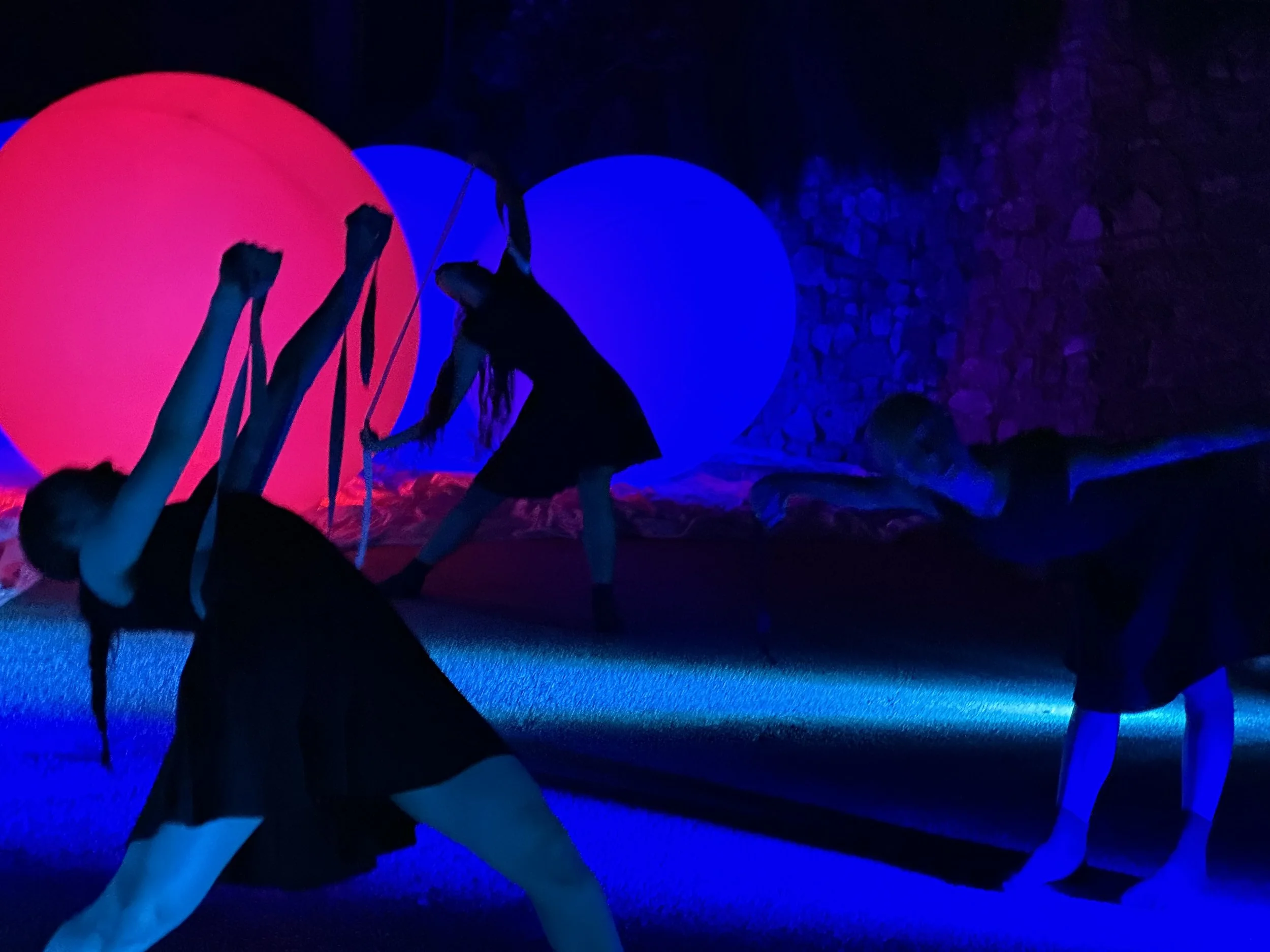 Bambini che giocano in un ambiente oscuro illuminato da luci al neon con sfondi di cerchi luminari e un muro di pietre.