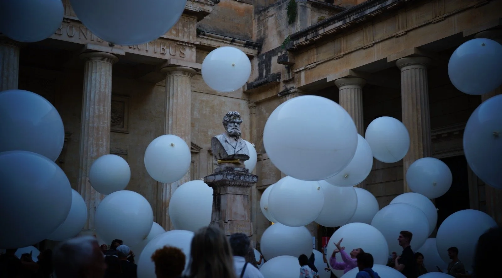 Scultura di un busto di un uomo con barba in un cortile antico circondato da numerosi palloncini bianchi e persone.
