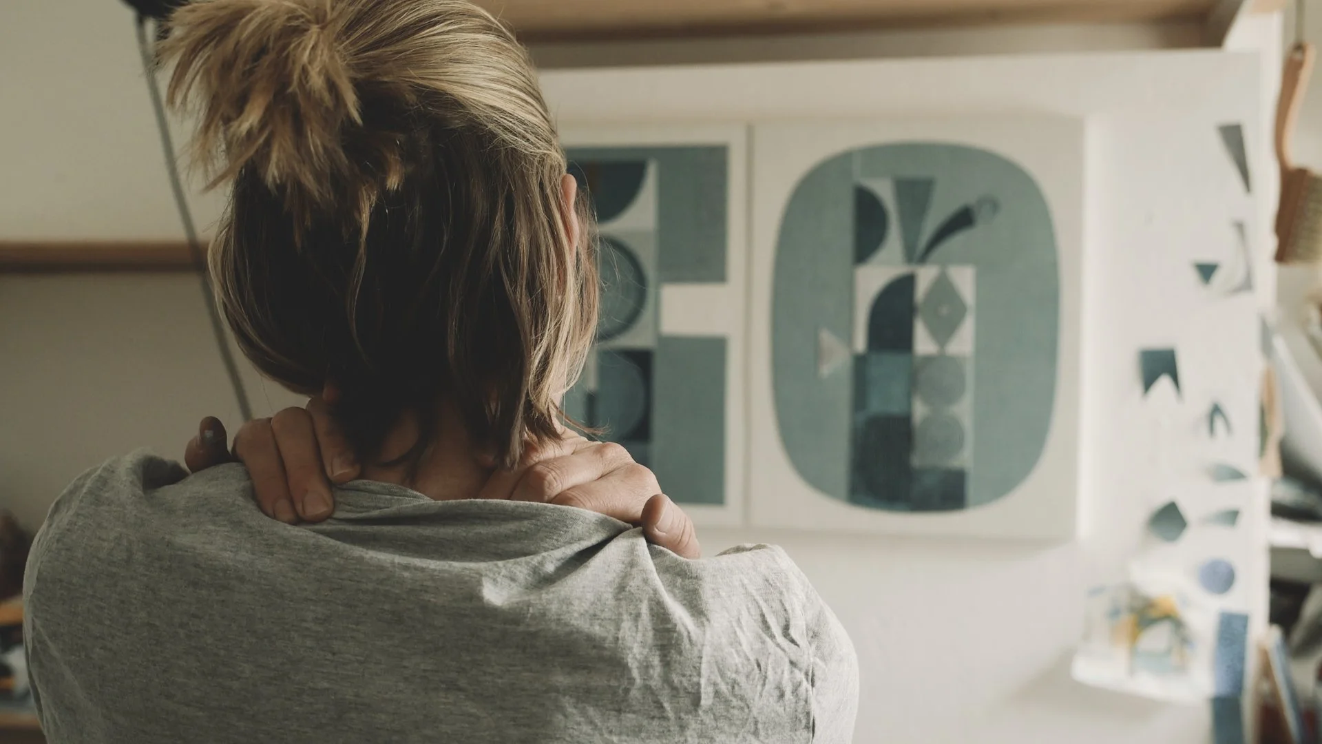 A person with short blonde hair wearing a gray shirt is hugging themselves with their arms crossed and hands resting on their shoulders in front of abstract artwork on a white wall.