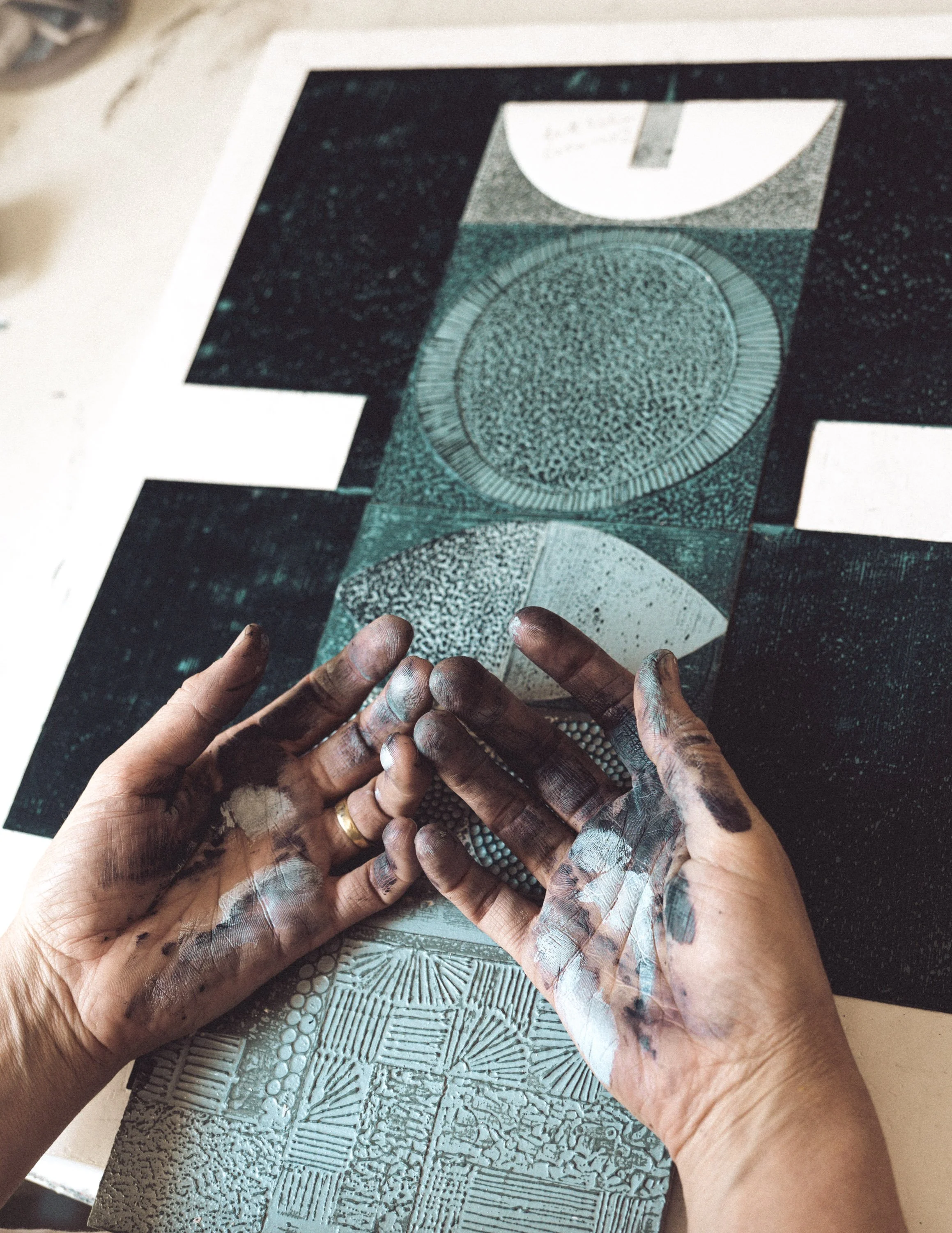 Hands covered in white and black paint working on a textured printing plate with geometric and circular patterns.