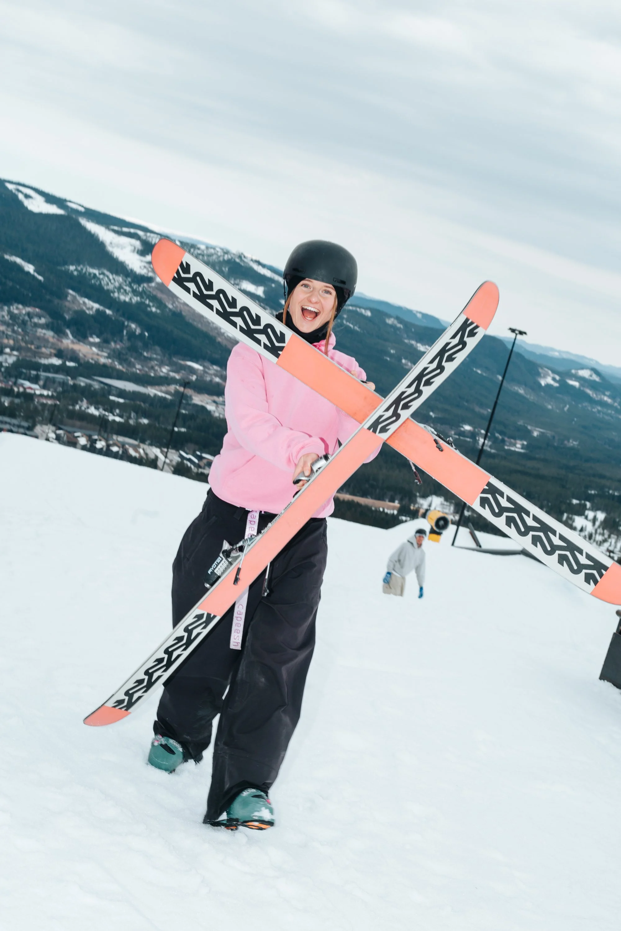 En kvinde i pink jakke og sorte snowboards bukser med hjelm og snowboards, smilende på ski i sneet landskab med bjerge i baggrunden.