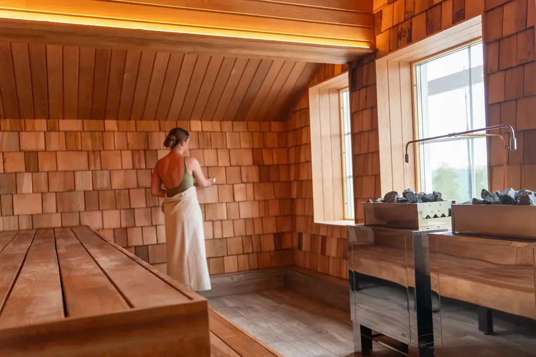 En kvinde står i en træsauna, set fra bagfra, med en hvid badetøj og håndklæde om livet, ved siden af to skorstene med varme sten