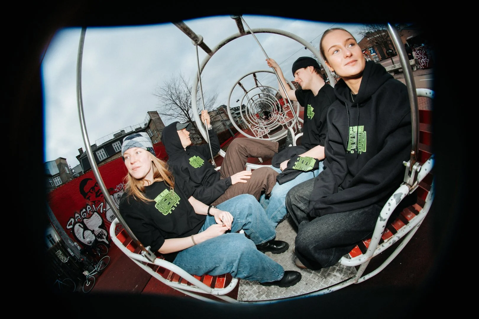 Four young people sitting on a spinning outdoor amusement ride, wearing black hoodies with green graphics, with a graffiti wall and bare trees in the background.