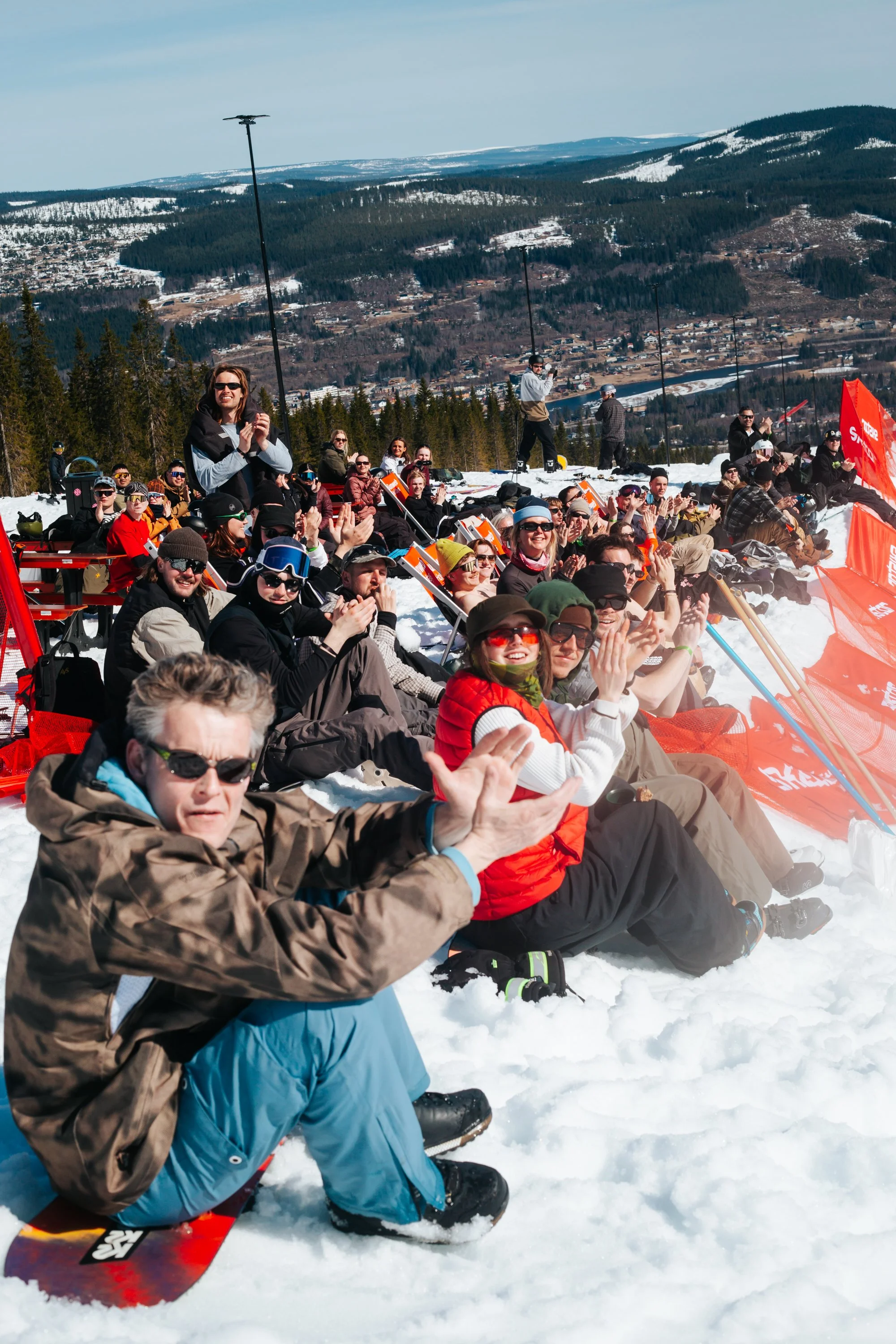 En gruppe mennesker sidder i sneen på ski i en bjerglandskab med skov og bjerge i baggrunden. Flere personer klapper og smiler, nogle har ski og snowboard, og der er orange banners.