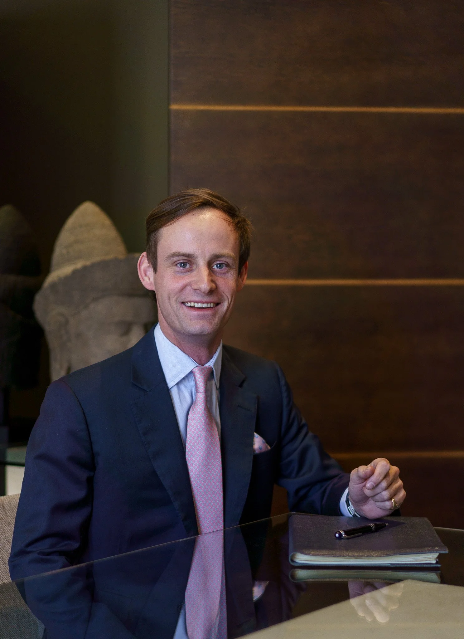 Man in business attire sitting at a glass table with a notepad and pen, smiling, with a stone sculpture in the background.