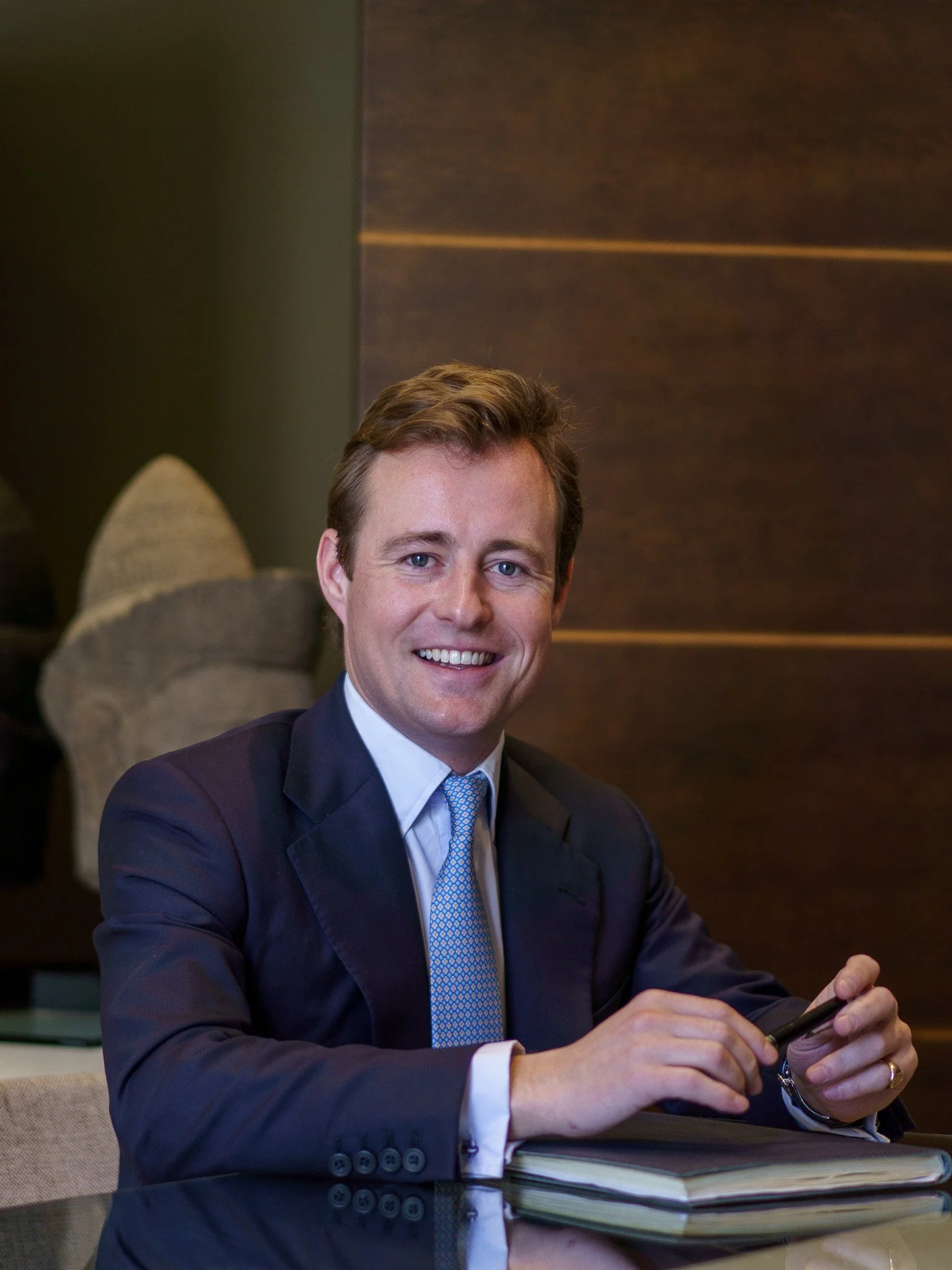 Smiling man in a suit holding a pen, sitting at a desk with a notebook.