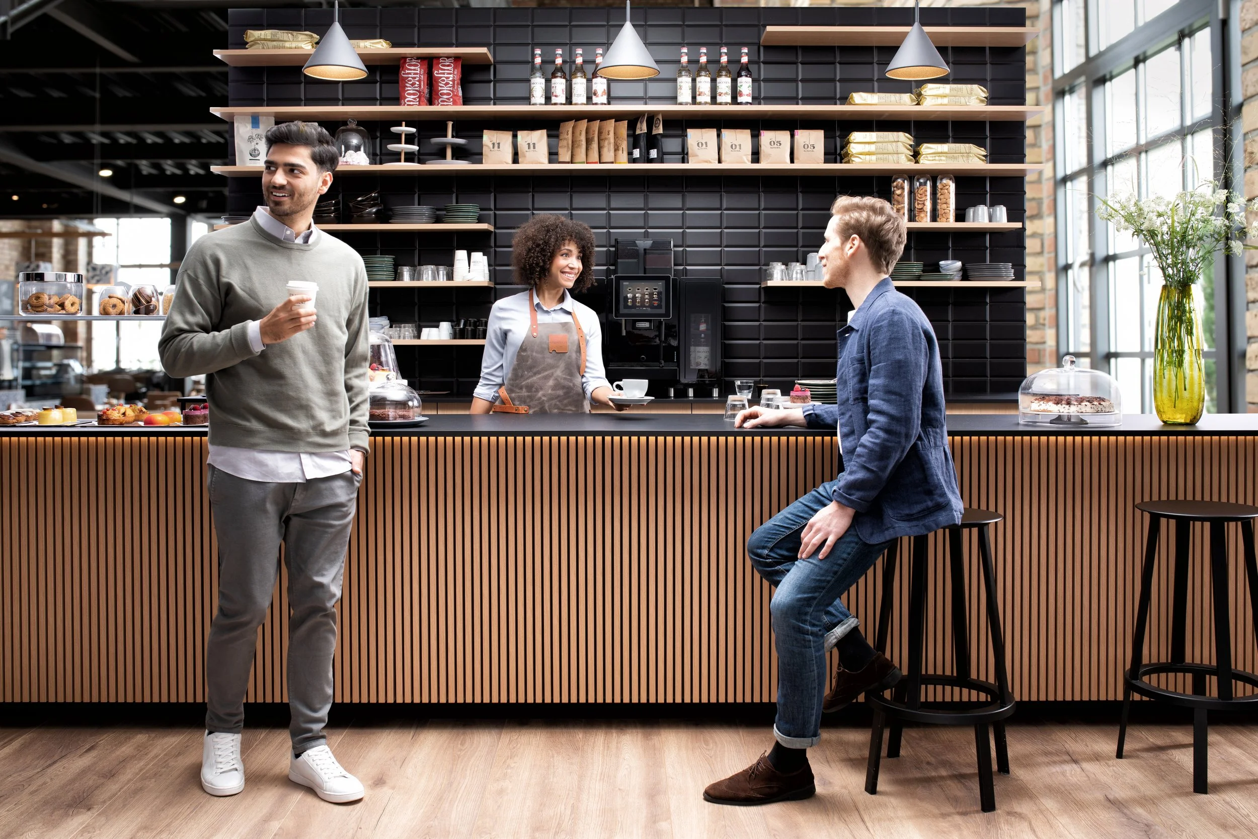 Two men talking at a coffee shop counter while a barista smiles behind the counter, which has pastries and a coffee machine, in a modern, well-lit cafe.