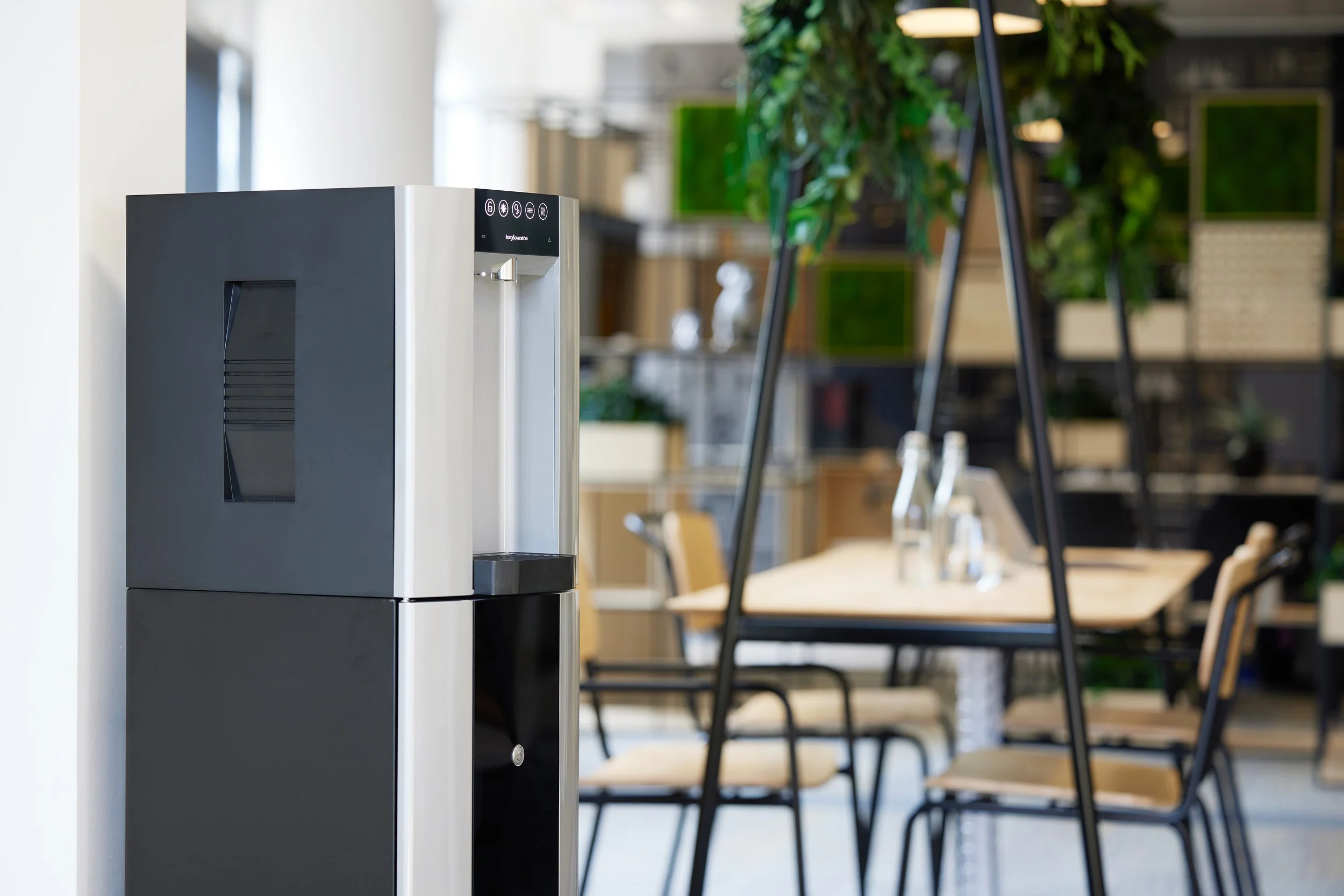 A modern water dispenser in a stylish office or cafe setting with wooden tables, chairs, glass bottles, and green plants in the background.