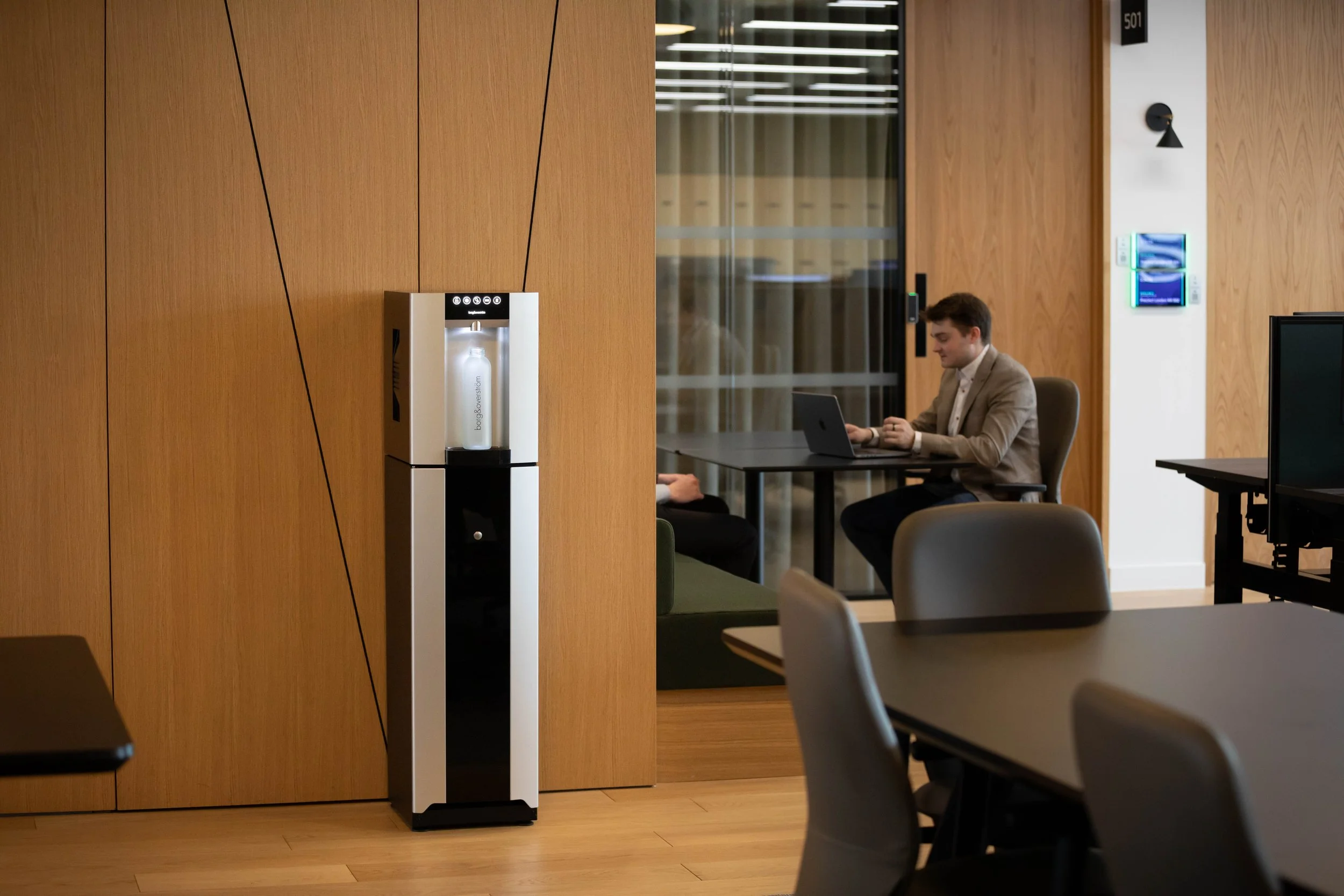 A man working on a laptop in a modern office conference room. A water cooler is positioned against a wooden wall near a glass partition, with tables and chairs in the foreground.