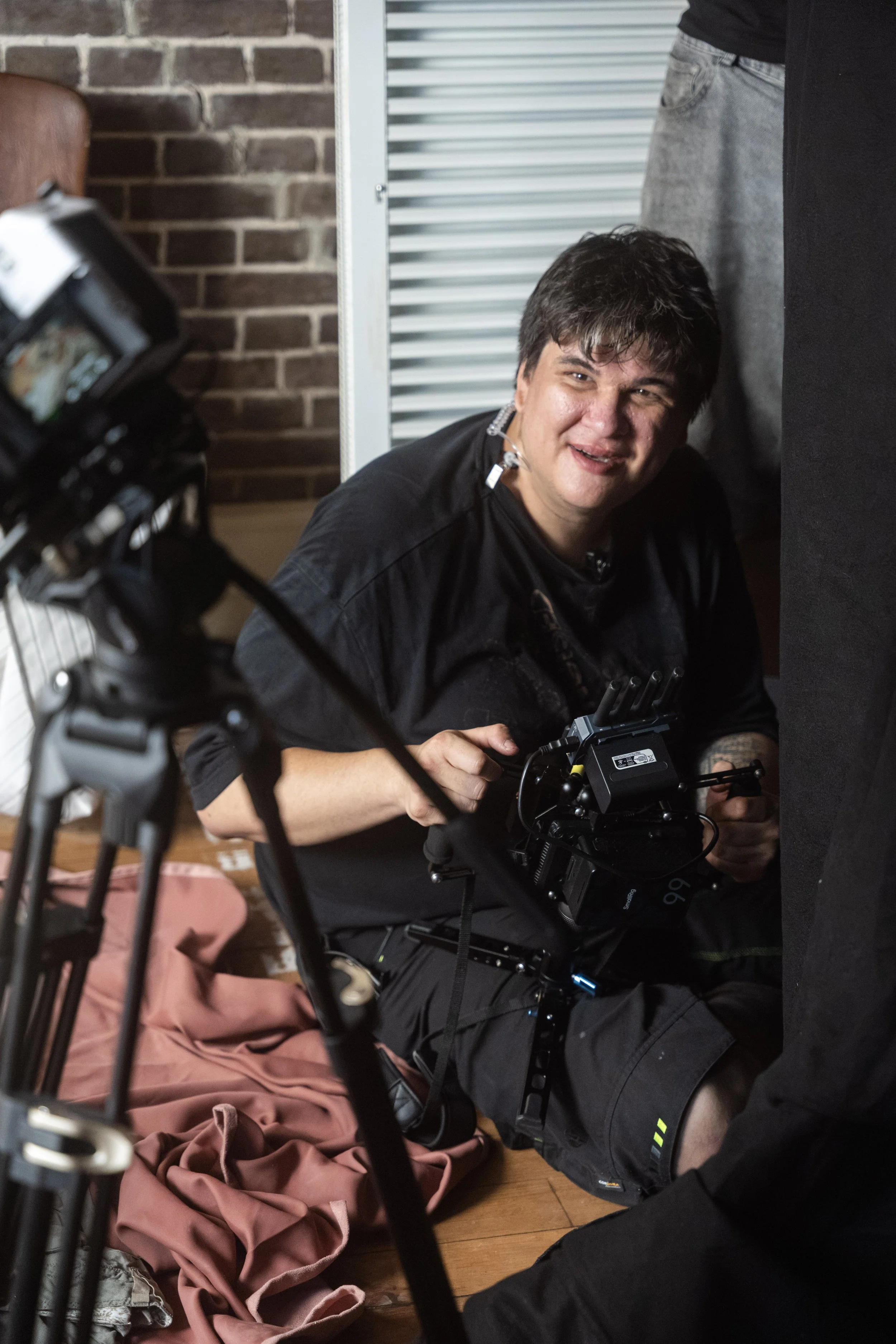 A man with dark hair and a black shirt is crouching on the floor, smiling, and holding some electronic equipment. There is a camera, a brick wall, and a metal radiator in the background, with a pink hoodie and camouflage shorts on the floor next to him.