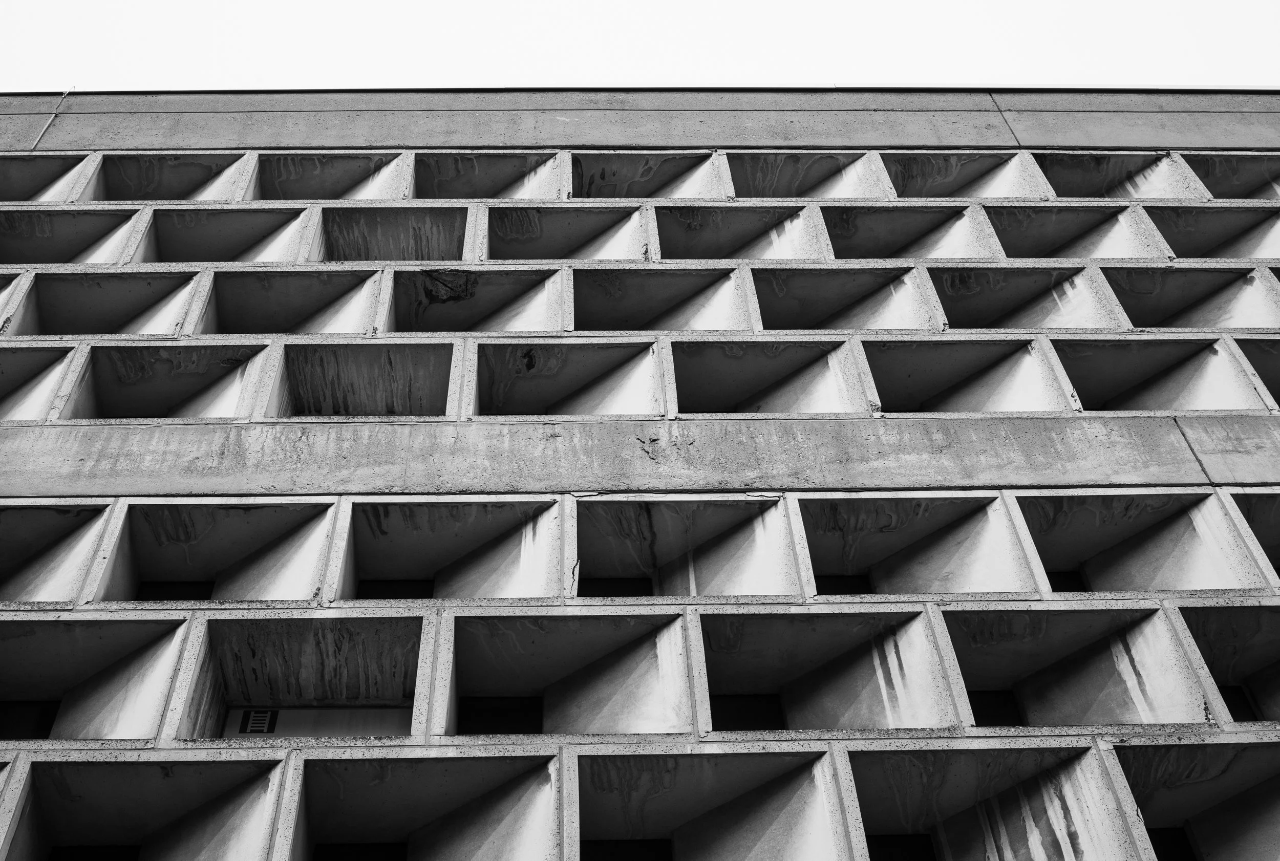 Black and white photo of a modern building's facade with a repetitive pattern of square concrete blocks and shadowed openings, viewed from below.