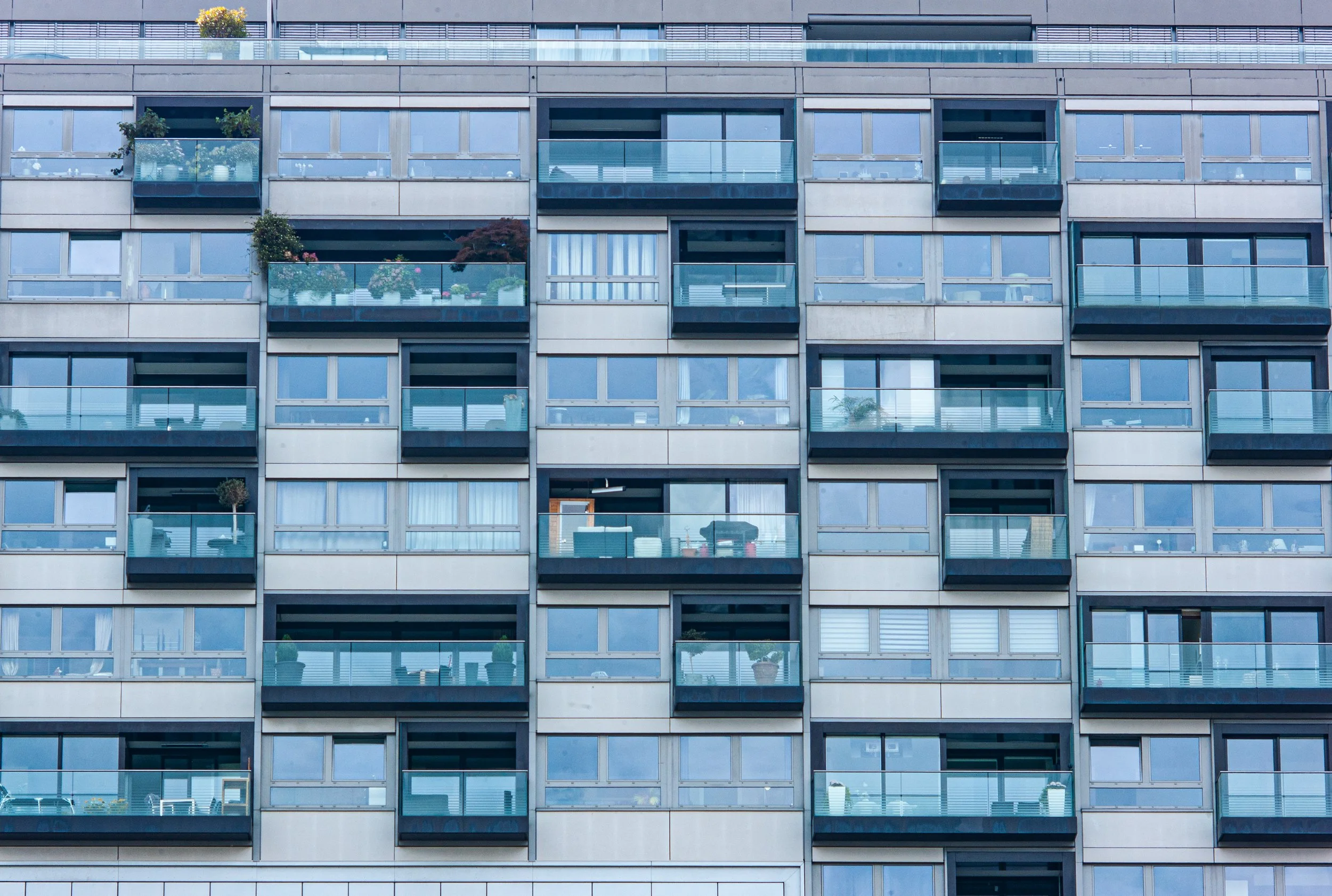 A multi-story apartment building with glass balconies and windows, some with potted plants and furniture visible.