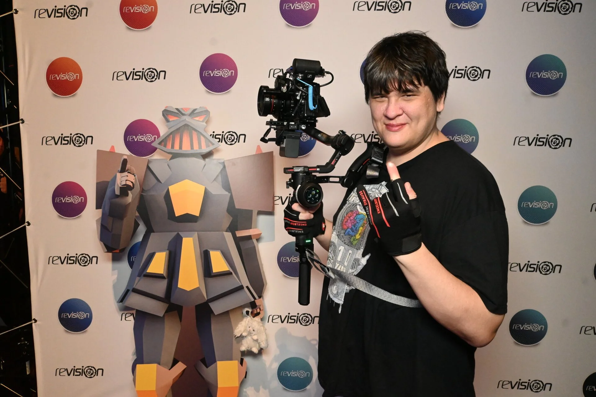 A young man with dark hair and a scruffy appearance smiling and giving a thumbs up at an event. He is holding a camera mounted on a stabilizer rig. The background features a step and repeat banner with the word 'revisión' repeated in a pattern along with colorful circular logos.