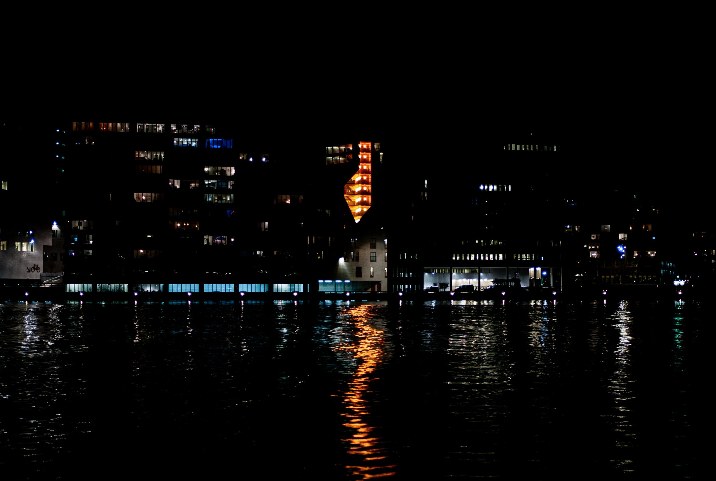 Night view of a city skyline over water, with illuminated windows and a building with orange lights.