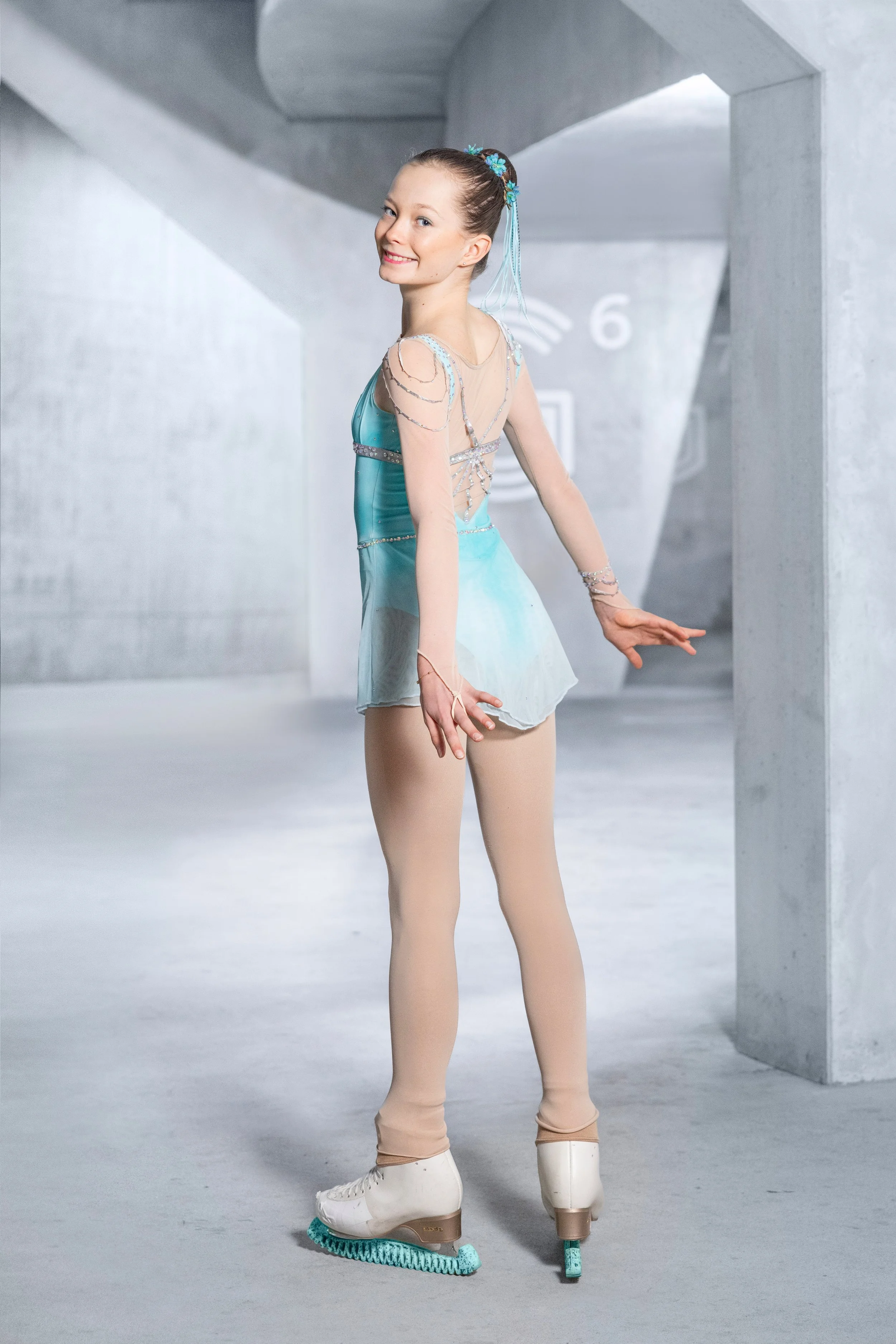 Eine junge Eiskunstläuferin in einem hellblauen Dress und Eishockey-Schuhen auf Kufen, die auf Eislaufbahn in einem modernen, grauen Gebäude zeigt.