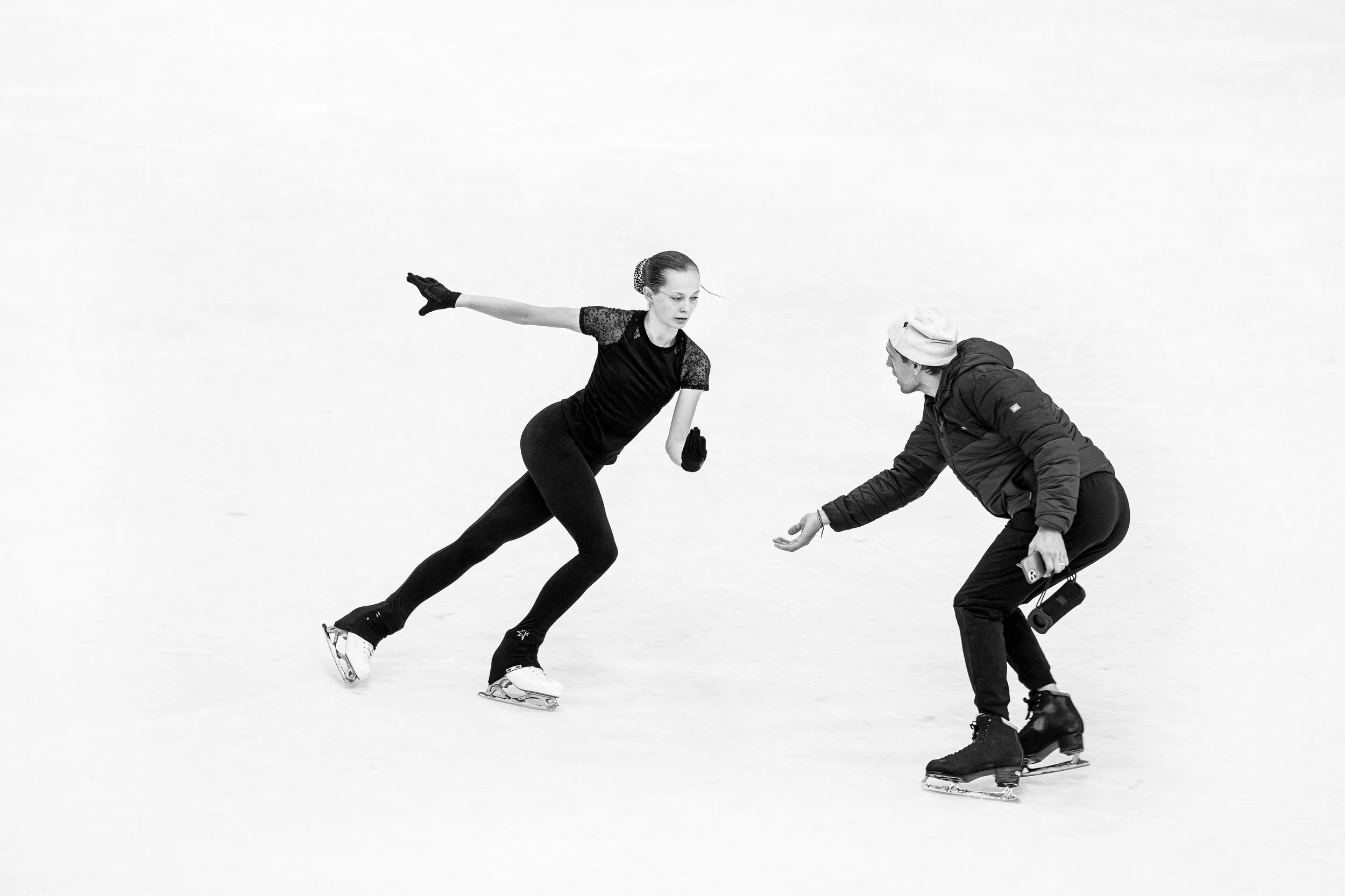 Eiskunstläuferinnen üben eine Szene auf dem Eis, eine trägt ein schwarzes Outfit mit einem glitzernden Haarband, die andere trägt einen dunklen Mantel und eine Mütze.