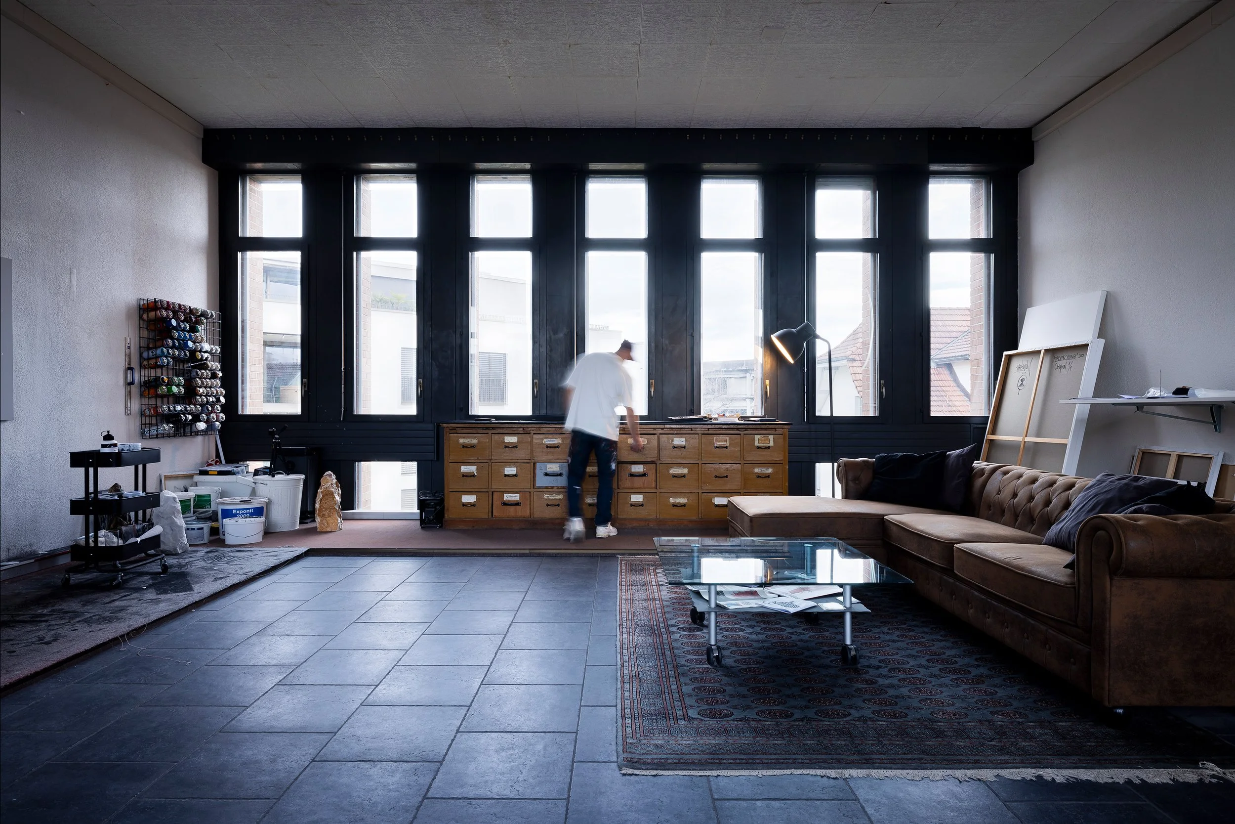 Ein Wohnzimmer mit großen Fenstern, einem braunen Ledersofa, einem Glastisch, einem Teppich und einer Person, die an einem Holzschrank arbeitet.