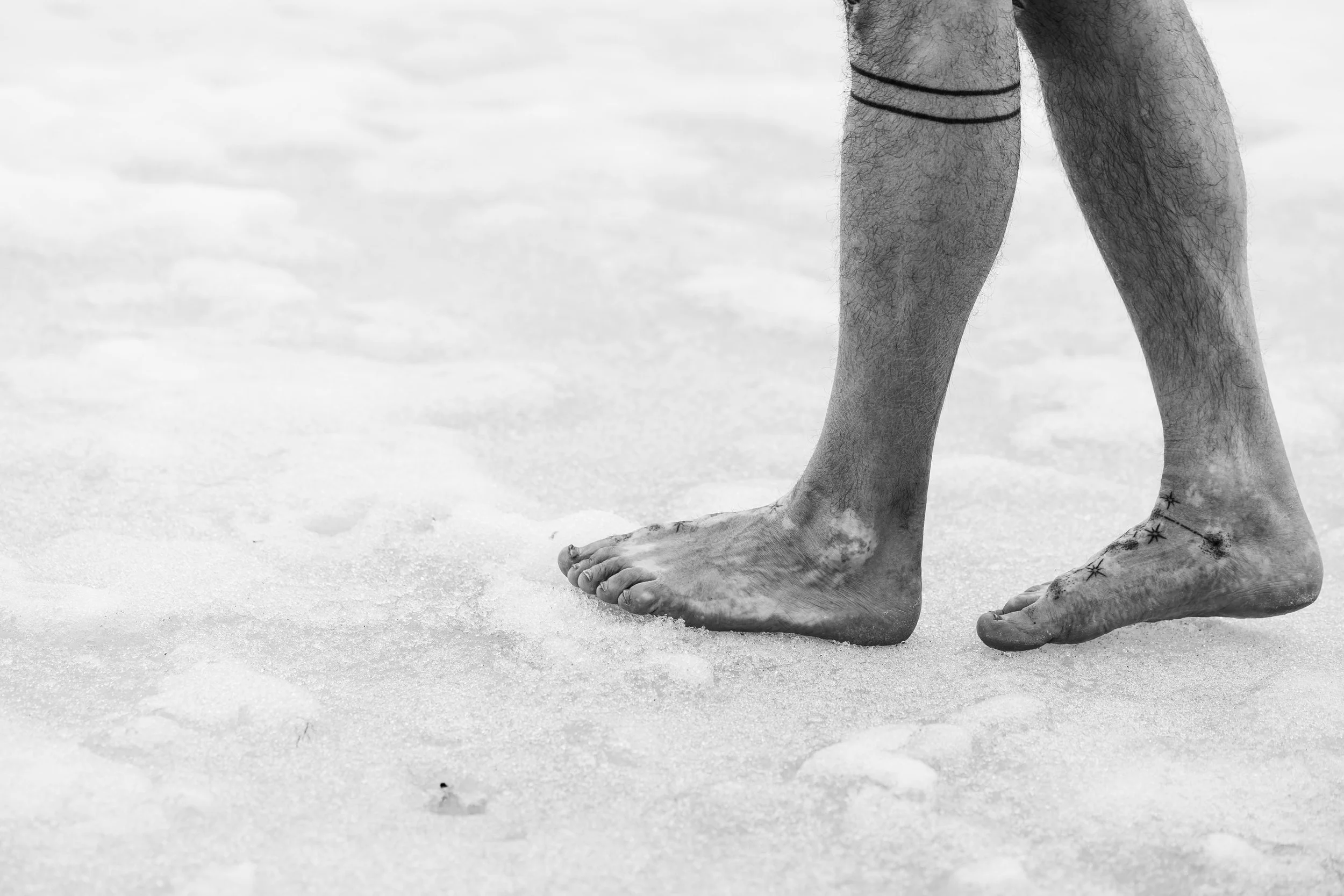 Ein menschliches Bein und Fuß auf Sand, mit Tattoos am Knöchel, im schwarzen und weißen Foto.